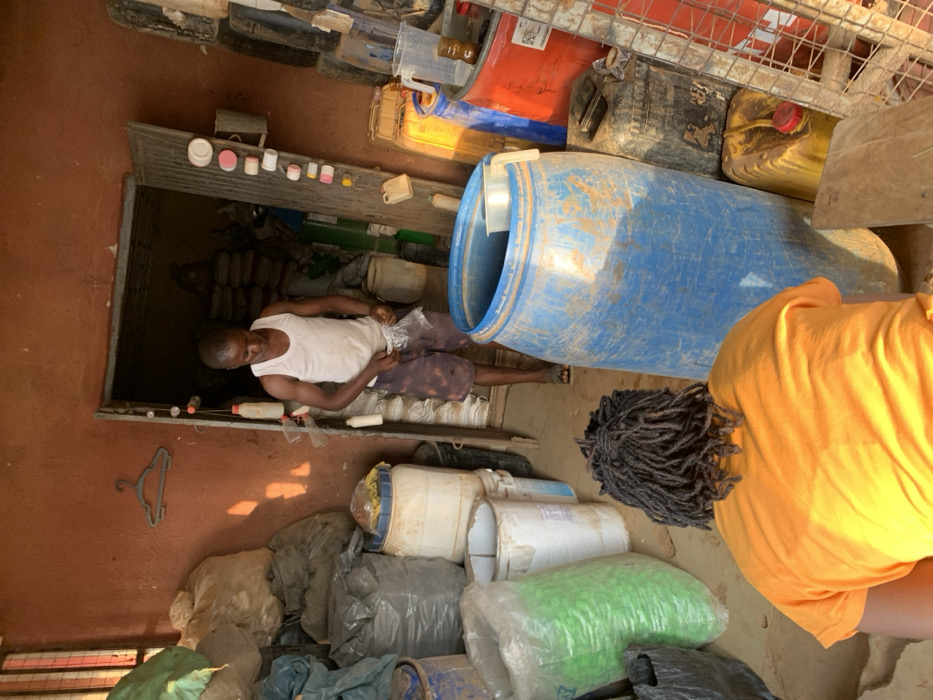 This image depicts an indoor scene in Ube, Nigeria, characterized by a utilitarian and cluttered environment. The reddish-brown walls suggest a non-residential structure, possibly a storage area or a workshop.
In the mid-ground, a male individual stands facing forward, positioned within a doorway that leads to a darker, equally cluttered room. He is wearing a white sleeveless tank top and dark-patterned shorts, with sandals on his feet. His hands are engaged with a clear plastic bag, which he appears to be opening or manipulating. To his left, mounted on the wall, is a dark grey plastic clothes hanger. Along the right side of the doorway frame, a string or wire holds several small, repurposed plastic bottles and containers of various colors, including white, pink, and red.
In the immediate foreground, partially obscured and with their back to the camera, is another individual. This person is wearing a short-sleeved orange shirt and has dark dreadlocks or braided hair. They are bending forward, their exact action not fully discernible.
The scene is dominated by numerous containers. To the left of the doorway, large plastic bags, some opaque grey, some filled with a light-colored substance, are stacked against the wall. A large green translucent bag is also visible on the floor, next to a white plastic barrel. To the right of the central standing figure, a large, cylindrical, weathered blue plastic barrel with an open top stands prominently; a white plastic pitcher or scoop is visible inside its rim. Surrounding this blue barrel are various other containers, including yellow and black jerrycans, a red barrel, and additional stacked tires or dark, cylindrical objects. A clear plastic pitcher with a handle is visible on top of one of the red containers on the right side.
Natural light appears to enter from an unseen source to the upper left, casting distinct shadows on the floor and objects. The overall impression is a busy, functional space dedicated to storage or processing of bulk materials.