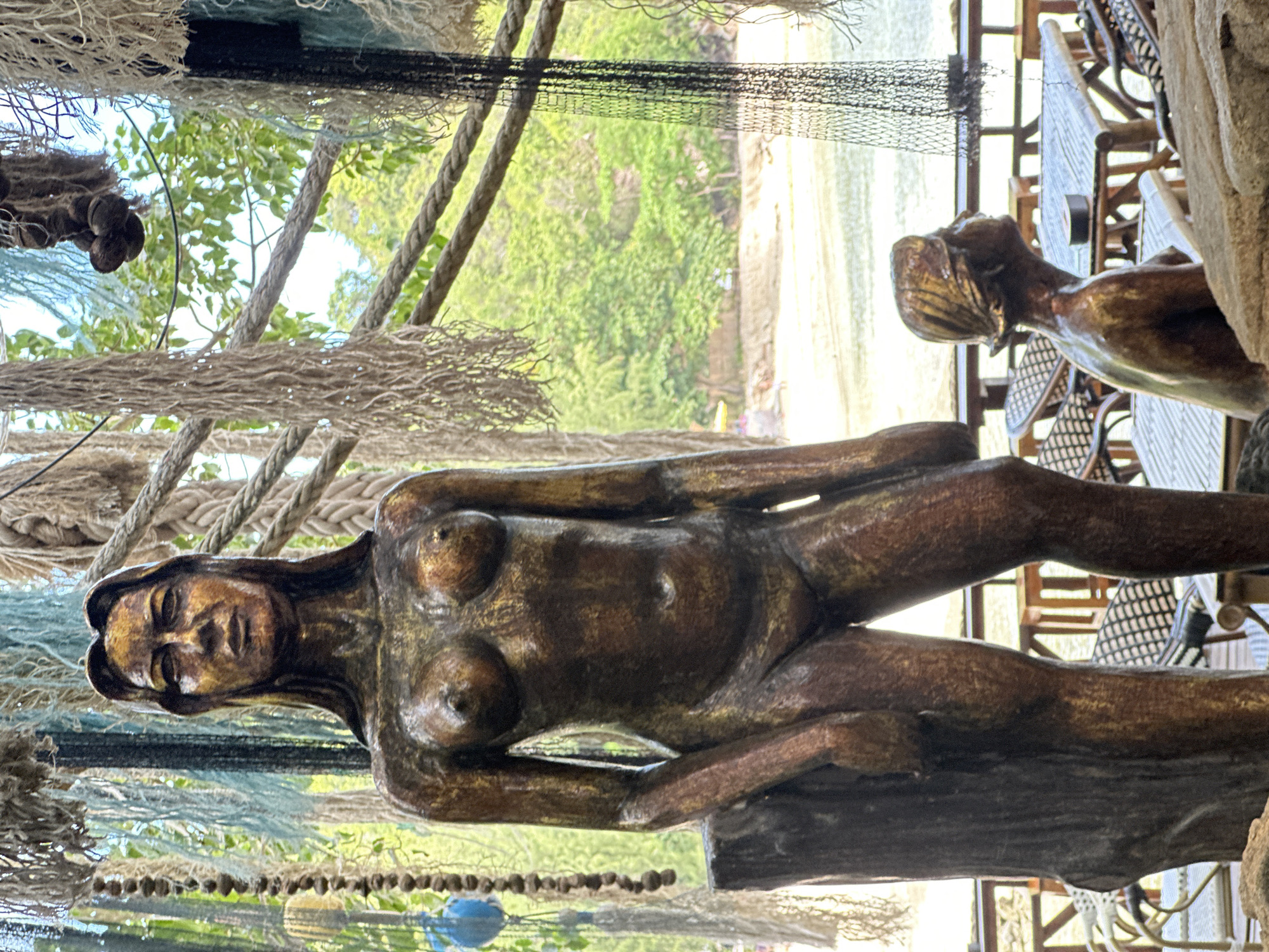 This media file depicts a bronze sculpture of a nude woman in a reclining pose. The sculpture is positioned in the foreground, with its head facing left and body angled towards the right. The sculpture's skin tone is a rich, metallic bronze, with visible detailing suggesting muscles and form. Her breasts are prominent, and her legs are bent, resting on what appears to be a dark, textured surface.
Behind the sculpture, a glimpse of an outdoor setting is visible. This includes greenery, likely trees and foliage, bathed in natural light. In the background, there are hints of tables and chairs, suggesting a restaurant or seating area, and a body of water, possibly the sea. The overall scene seems to be a well-lit outdoor environment.
The image has been rotated 90 degrees counter-clockwise, so the subjects are oriented sideways. This rotation affects the perception of the scene and the orientation of the sculpture. The time of day appears to be daytime, given the lighting conditions, and the weather seems pleasant and sunny.
The sculpture itself is the primary subject, with its detailed rendering drawing attention. The surrounding elements provide context, suggesting an artistic display or a decorative element in a tropical or seaside location. There is no visible text in the media file. The overall mood conveyed is one of artistic appreciation and a relaxed, outdoor atmosphere.