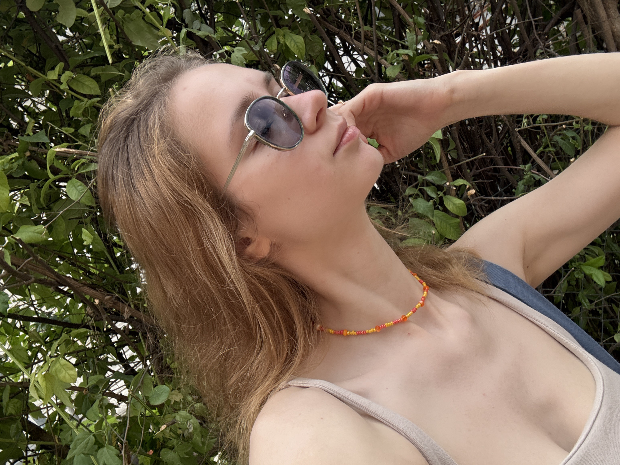 A young woman with light brown hair is resting her chin on her hand, looking down and to the side. She is wearing large, round sunglasses with a light colored frame and a beaded necklace in shades of orange and yellow. The background is filled with lush green foliage and branches, suggesting an outdoor setting. The woman is wearing a light-colored tank top, and part of a blue strap is visible. The scene appears to be taken in Kragujevac, Serbia, potentially in a park or garden area.