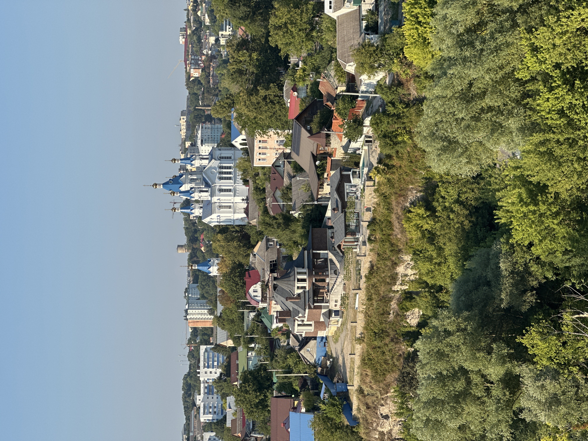 This is a high-angle, wide shot of the city of Kamianets-Podilskyi, Ukraine, on a clear, sunny day. The foreground is dominated by a dense canopy of lush green trees, with some lighter-colored foliage interspersed. A steep, rocky embankment rises behind the trees, topped by a line of stone fencing.
On the embankment and extending into the mid-ground are numerous residential buildings of varying architectural styles and sizes. Many of these are single-family homes with pitched roofs, some featuring dark roofing materials and brick or stucco facades. One prominent multi-story house with a conical turret and dark brickwork stands out in the mid-ground. There are also some smaller outbuildings and fences visible.
In the background, a large white church with multiple blue domes and golden spires is the most prominent structure. Adjacent to it, and further into the distance, are more urban buildings, including multi-story apartment blocks and industrial-looking structures. Cranes suggest ongoing construction. The sky is a clear, pale blue. The scene depicts a residential area within the city, nestled amongst natural greenery and overlooking a valley or ravine.