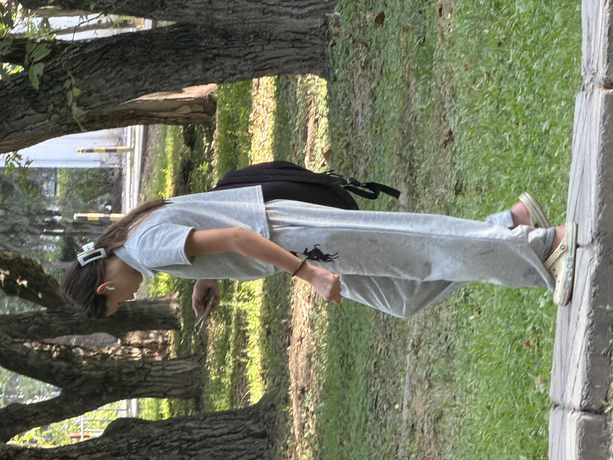 In an outdoor setting with trees and green grass, a young person with dark hair tied back with a white clip walks along a concrete path. They are wearing a grey t-shirt, grey sweatpants with a black horse emblem on the left leg, and light-colored clogs. The person is looking down at a smartphone they are holding in their right hand, and they have earbuds in their ears. A black backpack is slung over their left shoulder. The background shows more trees and some fencing.