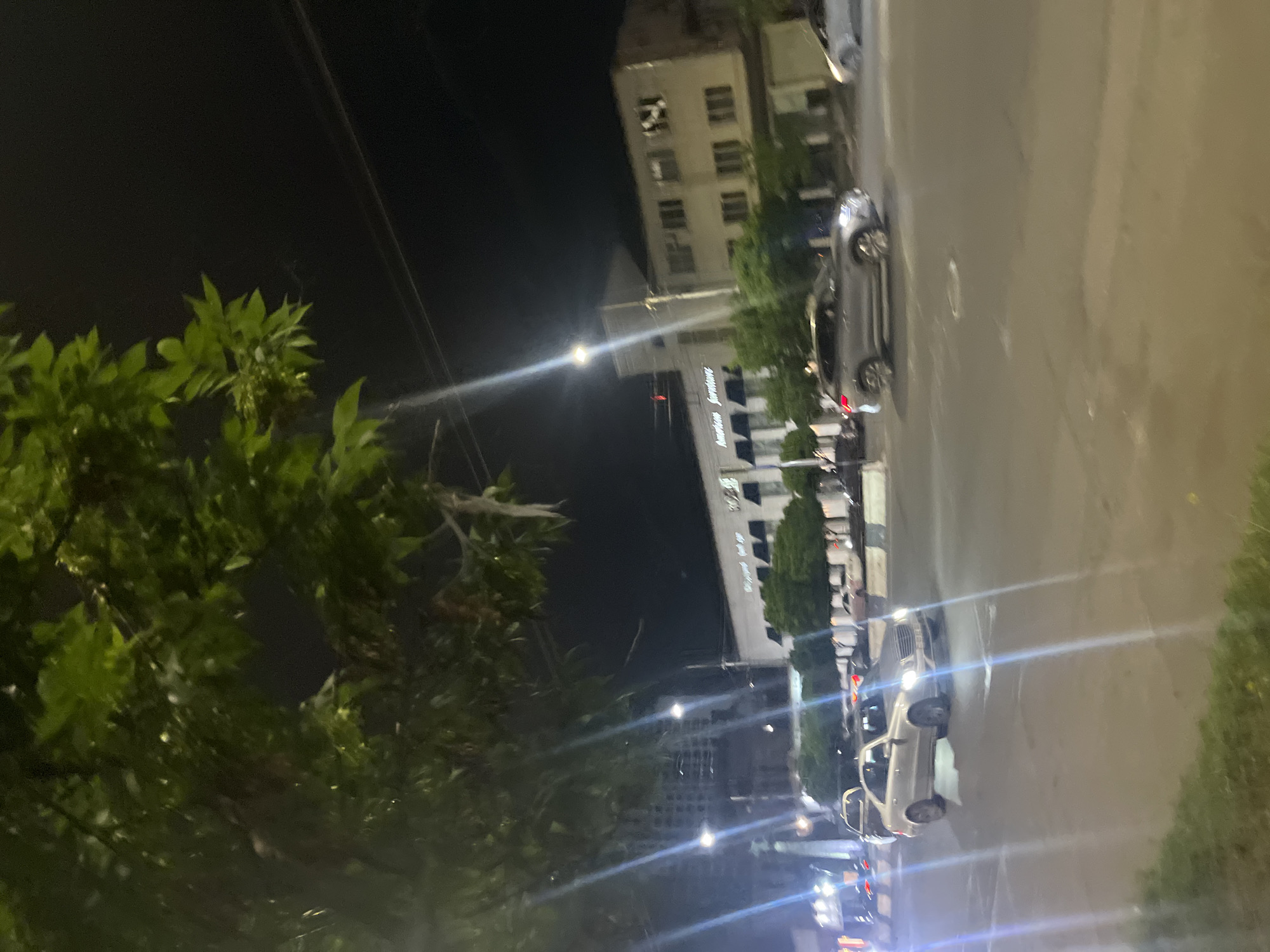 The image shows a night scene in Yerevan, Armenia. A street runs from the upper right to the lower left, lined with parked cars and buildings. "American Furniture" is visible on the side of one of the buildings. Trees occupy the left side of the frame. Streetlights illuminate the scene, causing lens flare. No people are distinctly visible.
