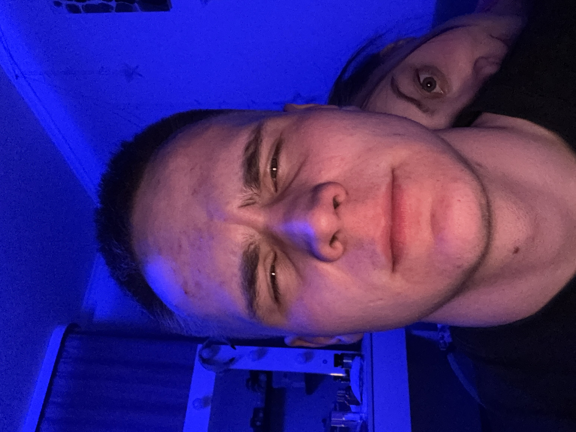 The image is a close-up, rotated 90 degrees clockwise, depicting two individuals in a dimly lit indoor setting dominated by intense blue lighting. A young person, likely male, is prominently featured in the foreground, facing the camera with a squinted, ambiguous expression that could be interpreted as intense, playful, or slightly uncomfortable. They have short, dark hair, and their skin appears reddish-pink under the blue illumination, with some visible blemishes on the forehead.
Behind and to the right of the foreground subject, another person is partially visible. Only one eye, wide open and looking directly at the camera, and a portion of their head are discernible, peeking from behind the main subject's shoulder or arm. Their hair also appears dark.
The background consists primarily of a solid, deep electric blue, indicating a wall or surface heavily illuminated by an artificial blue light source. Some faint textures or a small, dark rectangular object are subtly visible on the wall in the upper left corner. A dark-colored object, possibly part of a furniture item or equipment, is also partially visible behind the foreground person's head. The strong blue light creates a dramatic and somewhat ethereal atmosphere, suggesting the photograph was taken in a low-light environment, likely at night or in the evening. No visible text is present in the image.