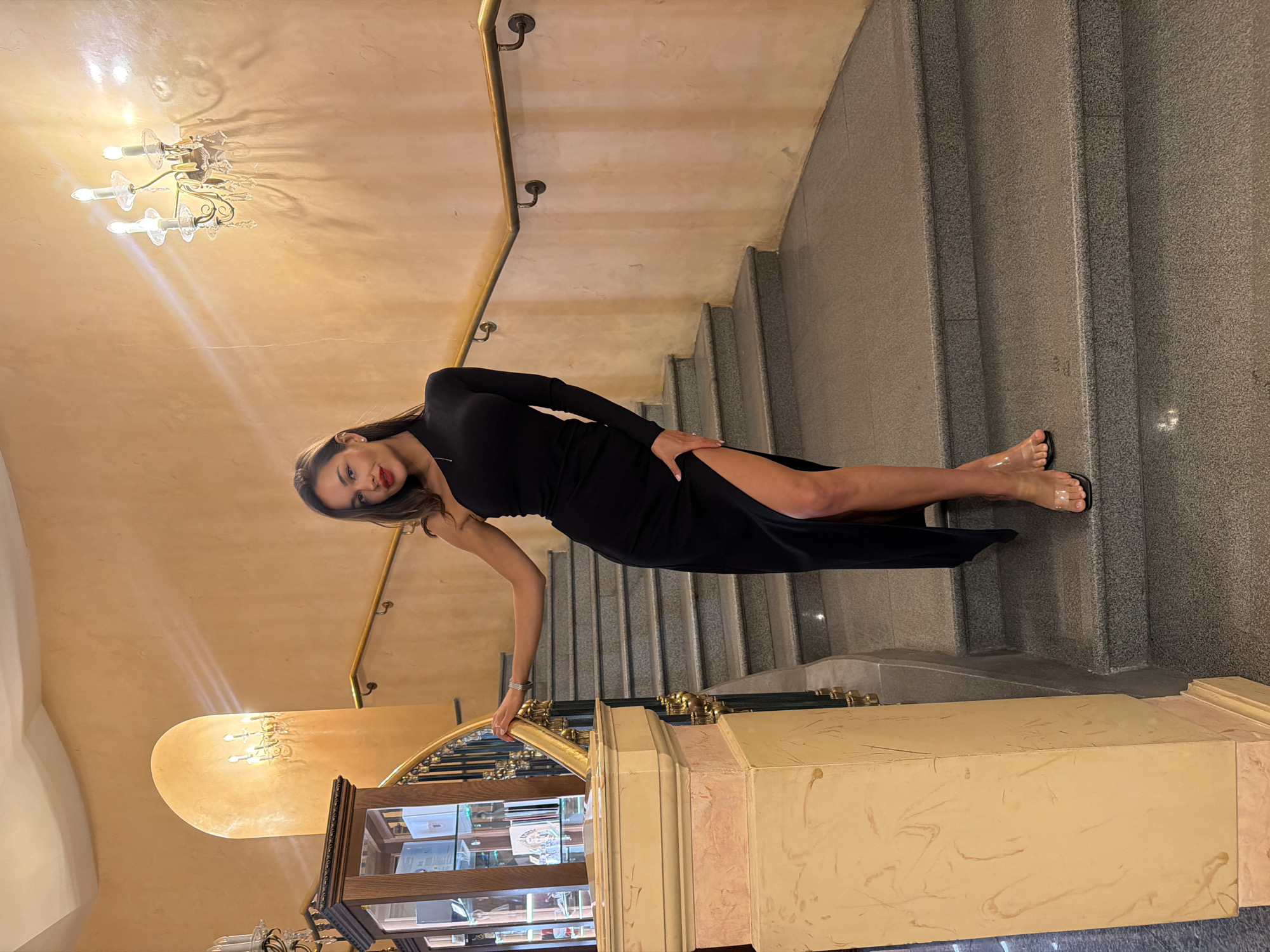 An elegantly dressed woman poses gracefully on a grand staircase within a luxurious indoor setting, likely in Moscow, Russia. The scene appears to be set in the evening or night, illuminated by warm artificial light.
The central figure is a woman with long dark hair, wearing a sleek, long-sleeved black dress that features a high side slit, revealing her leg. She is wearing red lipstick and clear-strapped sandals. Her left arm is bent, with her hand gently resting on a polished brass handrail, while her right hand is casually placed on her hip. She maintains eye contact with the camera, conveying a confident and poised demeanor.
The interior boasts a wide, sweeping staircase constructed from grey, speckled granite or marble, with distinct steps leading upwards. The walls are a warm, textured beige or light brown, subtly reflecting the light. Ornate, classical wall sconces with multiple candle-like bulbs cast a golden glow, creating atmospheric shadows on the wall surfaces. Below the staircase, a substantial decorative pillar with intricate patterns in shades of beige and light pink is visible, along with a wooden display case containing various items. The overall impression is one of opulence and refined architecture. No visible text is present in the image.