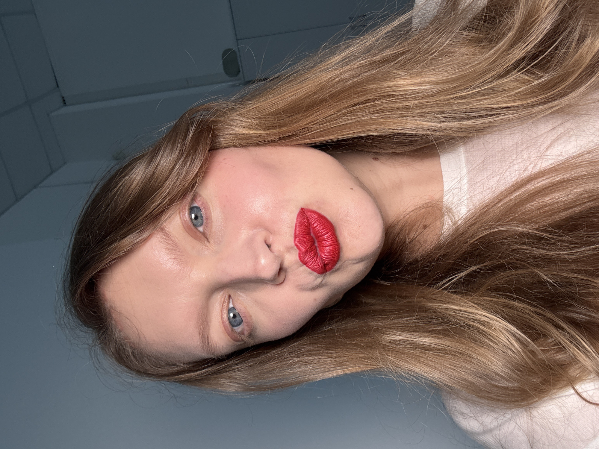 A head-and-shoulders portrait depicts a fair-skinned female, estimated to be in her 20s or 30s, looking directly into the camera. She has long, wavy, light brown hair with lighter highlights, parted on her left, cascading around her face and over her shoulders. Her eyes are blue. She is wearing a white, crew-neck garment. Her lips are adorned with vibrant, matte red lipstick and are pursed into a puckered, "kissing" expression. Subtle pink blush is visible on her cheeks. A small brown mole is present on her lower left cheek, and another on the right side of her neck, just below her jawline. The background features a plain, cool blue-grey wall on the left. On the right, architectural elements of an indoor environment are visible, including a darker ceiling section with a fixture or vent, and a white partition or door frame. The subject is illuminated by direct frontal lighting. This scene is located in the city of Izhevsk, Russia.
