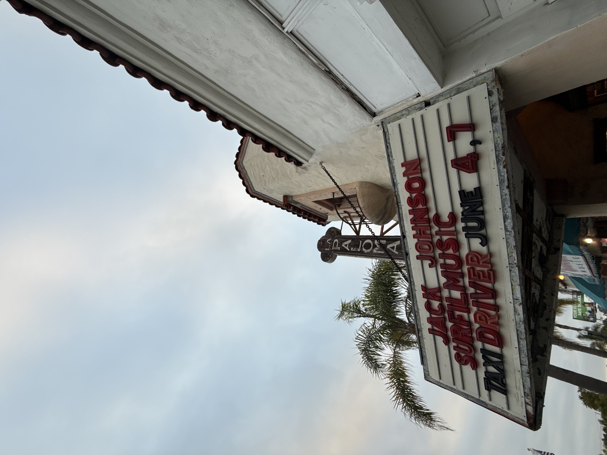 An upward-angled view captures the La Paloma Theatre in Encinitas, California, United States of America. The main focus is the theatre's marquee and the upper portion of its Spanish Colonial Revival-style building.
The marquee, a white slatted sign with a metal frame, displays red and black block letters. The top line reads "JACK JOHNSON" in red. The second line states "SURF FILM MUSIC" also in red. The bottom line, "TAXI DRIVER JUNE 4, 7," uses black letters for "TAXI DRIVER" and red letters for "JUNE 4, 7". The marquee shows signs of weathering and minor rust along its lower edge.
Above the marquee, a vertical sign with a decorative finial spells "LA PALOMA" downwards. The building itself features textured white stucco walls and a distinctive red barrel-tile roof. A small, decorative balcony with a light-colored stucco base, an ornate iron railing, and a chain detail is visible on the upper left side of the structure.
Several palm trees frame the theatre, with their fronds extending into the upper left and lower right portions of the image. The sky is a uniform, soft blue-grey, indicating an overcast condition or late afternoon light. No individuals are visible in the frame. A small green awning and a partial sign for an adjacent business are subtly visible at the bottom of the image.