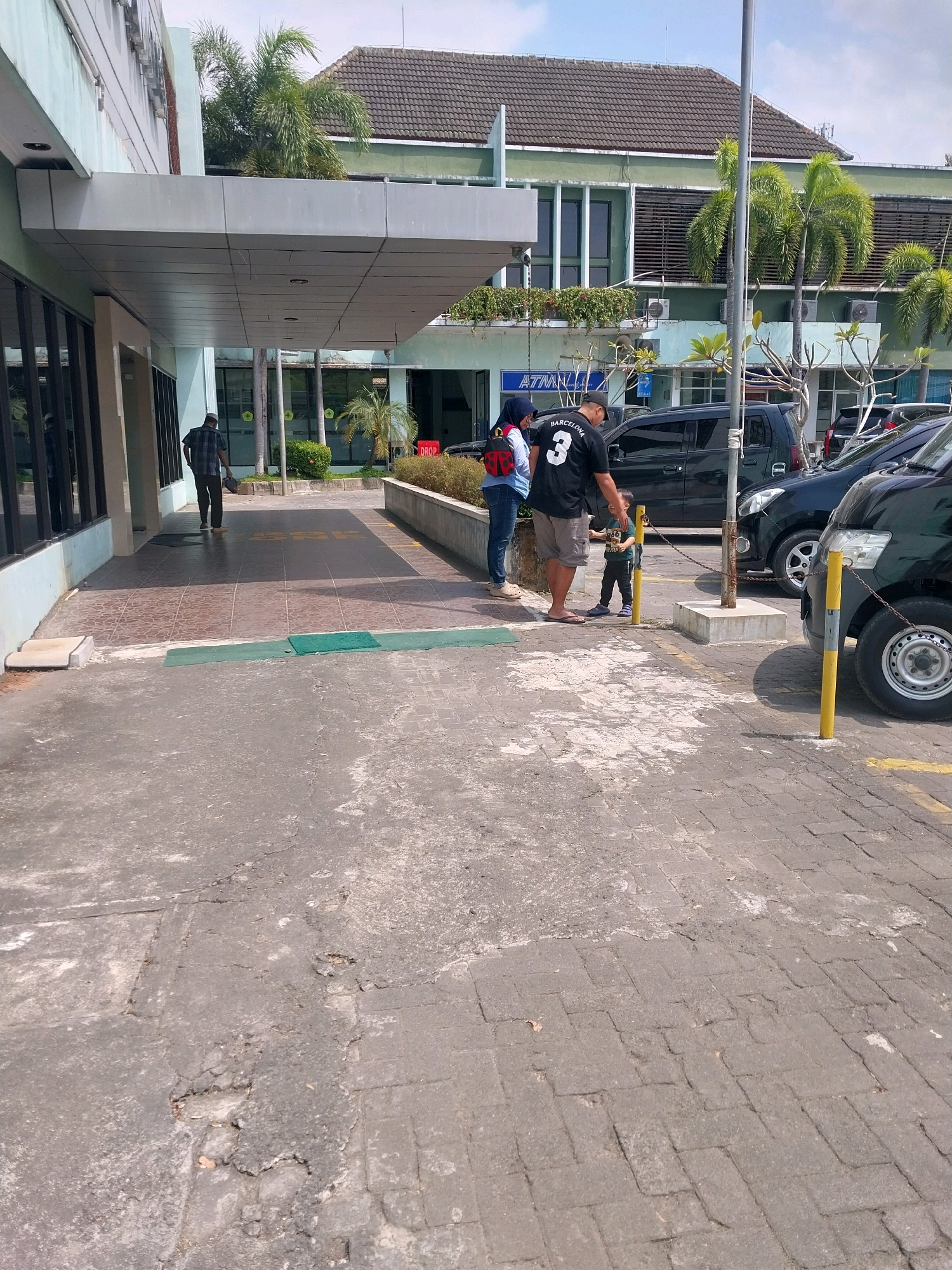 In Wates, Indonesia, a paved outdoor area features a building with a covered walkway. A man in a plaid shirt is sweeping the tiled walkway, while a woman in a hijab and jeans stands with a young boy wearing a black shirt and shorts. The woman has a red backpack on her shoulder, and the boy is holding a small object. Behind them, several dark-colored vehicles are parked. Palm trees and other greenery are visible in front of the building, which appears to be an office or commercial establishment with the sign "AZMAN" visible. The ground is a mix of cracked concrete and interlocking pavers.