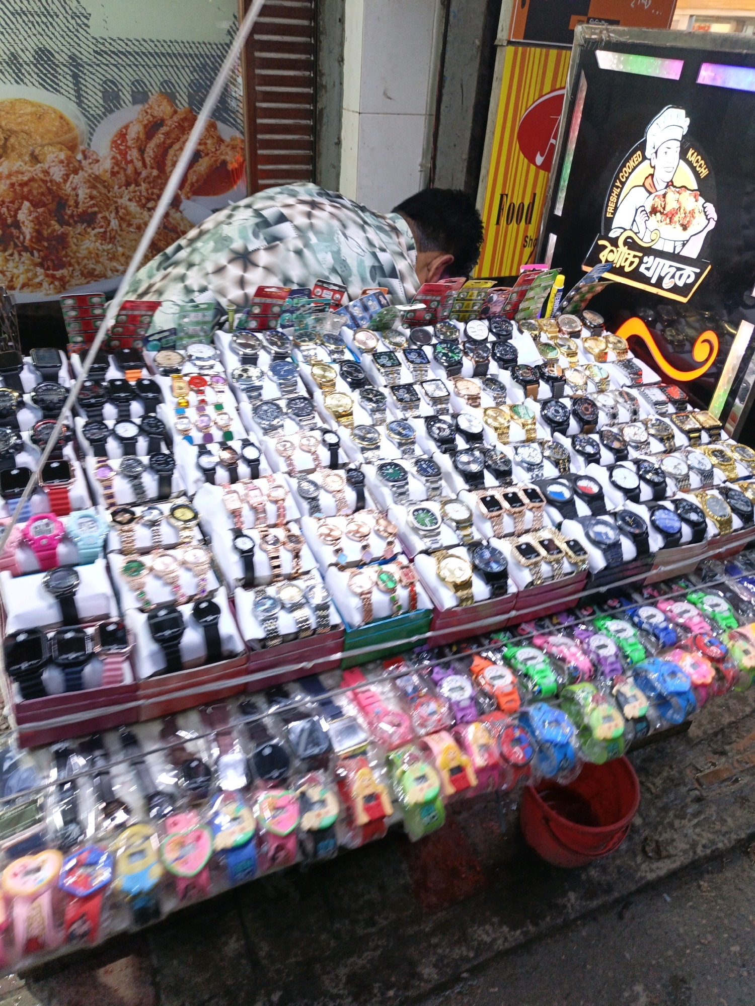 The image depicts a street-side stall in Dhaka, Bangladesh, where a vendor is selling a variety of watches. The stall is laden with numerous watches arranged in display boxes, showcasing a wide range of styles, from classic metal bands to colorful, novelty designs, including cartoon-themed watches. In the background, a sign for "Kacchi Biryani" and "Freshly Cooked" suggests a food establishment, possibly indicating the stall is located in a bustling commercial area. A person, likely the vendor, is partially visible in the background, tending to the stall. The setting appears to be an outdoor market or sidewalk, with a wet, uneven ground suggesting recent rain or a generally damp environment. The overall scene captures the vibrant street commerce typical of Dhaka.
