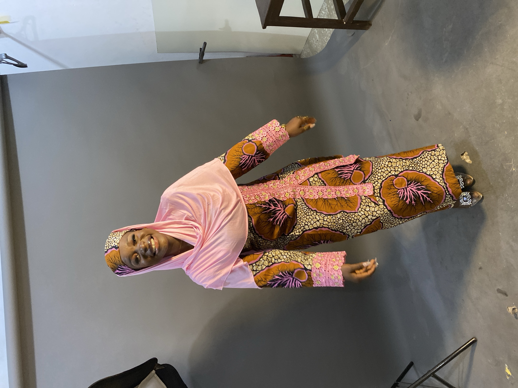 A portrait photograph captures a smiling woman standing and posing indoors, in what appears to be a photography studio. The woman is the central subject, looking directly at the camera with a broad, joyful smile. She is dressed in a vibrant, floor-length gown featuring a distinctive pattern of large, abstract shapes in shades of brown, black, and pink. The dress is adorned with intricate pink lace or embroidery along the neckline and cuffs, interspersed with small golden accents. A soft pink headscarf is draped over her head and shoulders, complementing the pink elements of her dress. Her feet are visible, wearing open-toed shoes or sandals embellished with a sparkly silver ornament.
The setting is simple, with a plain grey wall forming the background and a matching grey floor. The upper part of the background transitions to a lighter, possibly white or off-white, area, suggesting a studio setup. Minimal objects are visible in the periphery: a metal clothing hanger is on the wall in the upper left, and a partial view of a dark, metallic stand or chair leg is on the floor in the lower right. The overall lighting is bright and even, consistent with an indoor photoshoot. Based on the provided context, the location is Gura Top, Nigeria. There is no visible text in the image.