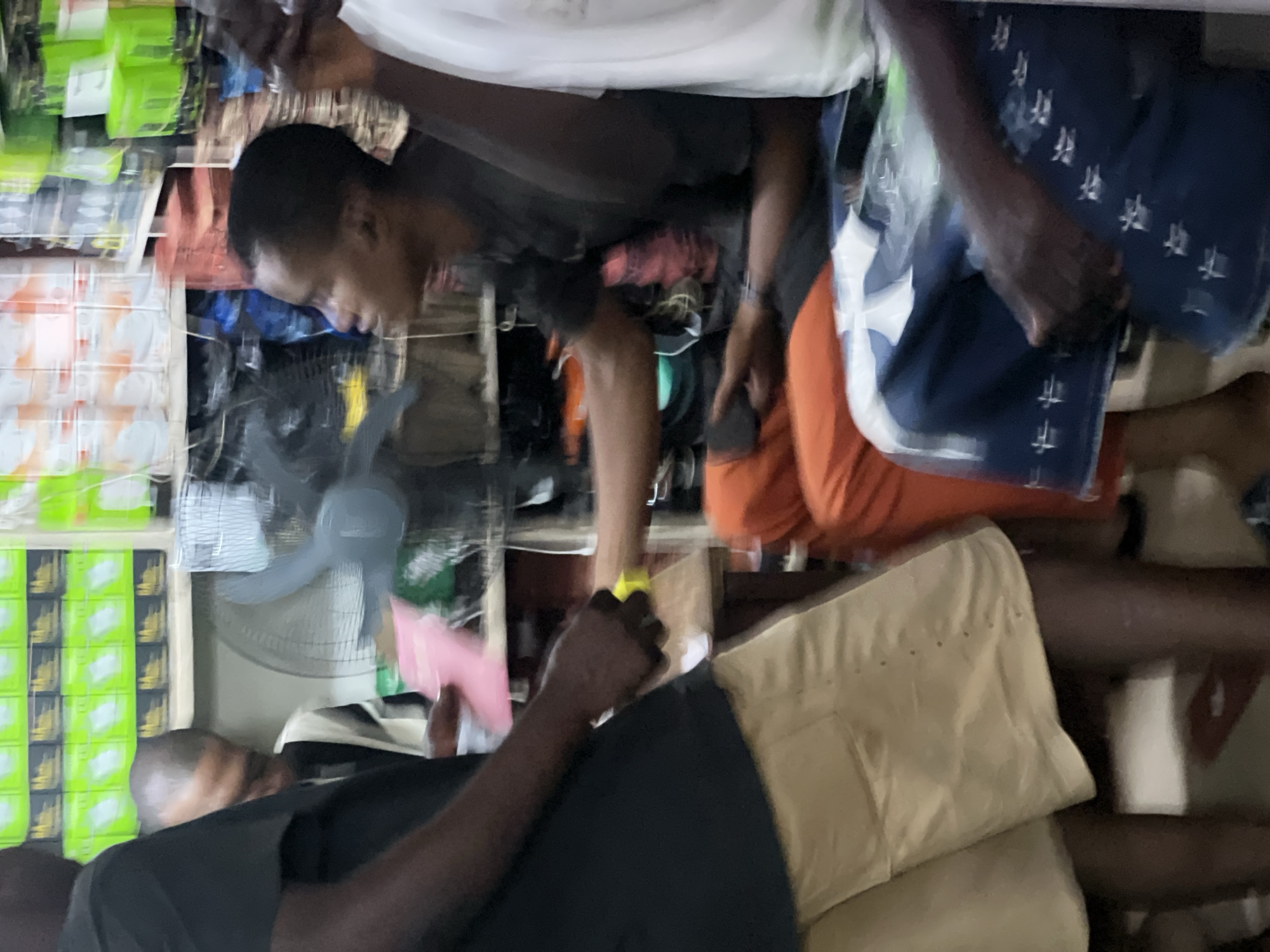In a shop at Idimu, Nigeria, two individuals are present. One individual wearing an orange garment and a blue garment with white patterns is standing behind another. The other person is wearing a black garment and tan shorts, and a yellow object is in their hand. An oscillating fan is visible in the background along with shelving filled with assorted items.