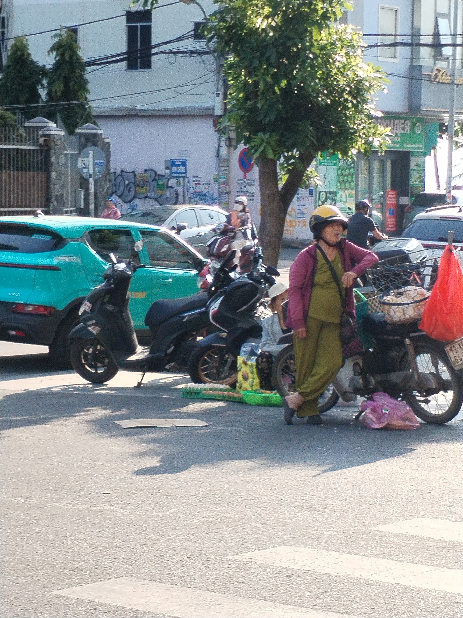 The image shows a street scene in Bac Nha Trang Ward, Vietnam. A middle-aged woman wearing a yellow helmet, olive green outfit, and a pink cardigan stands near a parked motorcycle loaded with goods. She appears to be leaning on the motorcycle. A scooter and a turquoise car are parked nearby. In the background, there are buildings with graffiti and shop signs in a foreign language. Other people and vehicles are visible in the background, indicating a busy street environment. A crosswalk is apparent in the foreground.