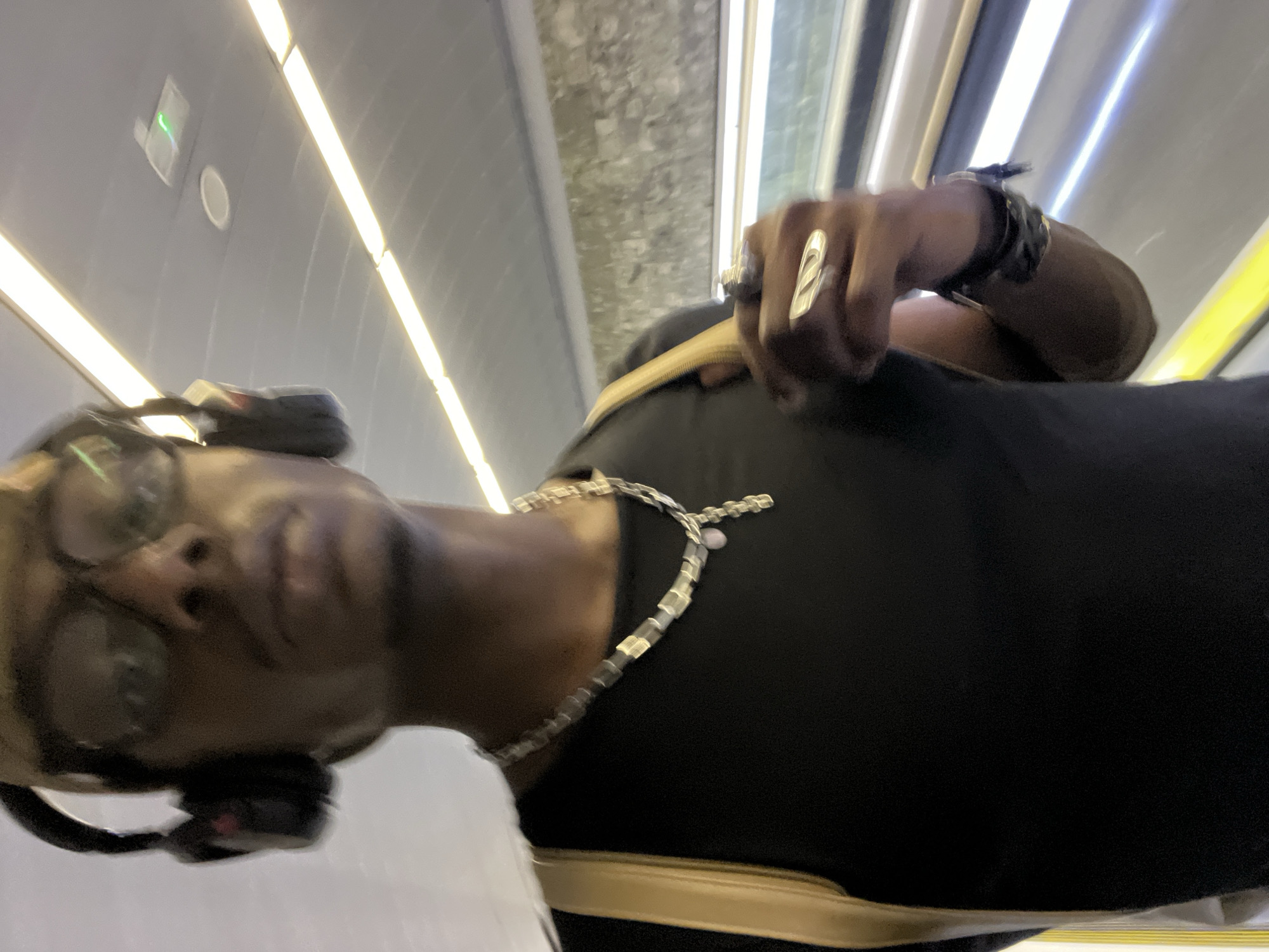 A vertically oriented image depicts a person, a male with a dark skin tone, inside what appears to be a modern train or metro car, potentially in Barcelona, Spain. The person is positioned in the lower left of the frame, looking towards the camera with a relaxed, slightly smiling expression. He wears clear-framed glasses and black headphones are resting around his neck or chin. A silver-colored chain necklace with rectangular links is visible over his black t-shirt. On his right hand, a distinctive silver-toned ring with an abstract, swirling design is prominent, along with a dark-colored watch or bracelet on his wrist. A beige or tan strap, likely from a bag or garment, is seen draped over his shoulder.
The background reveals the interior of the vehicle, characterized by a curved, light-colored ceiling. Multiple bright, linear light fixtures run along the ceiling, providing artificial illumination. A faint reflection on the ceiling shows a small green light indicator and a circular vent or speaker. A window or a bright light source from outside the vehicle is visible on the right side of the frame. No discernible text is visible in the image.