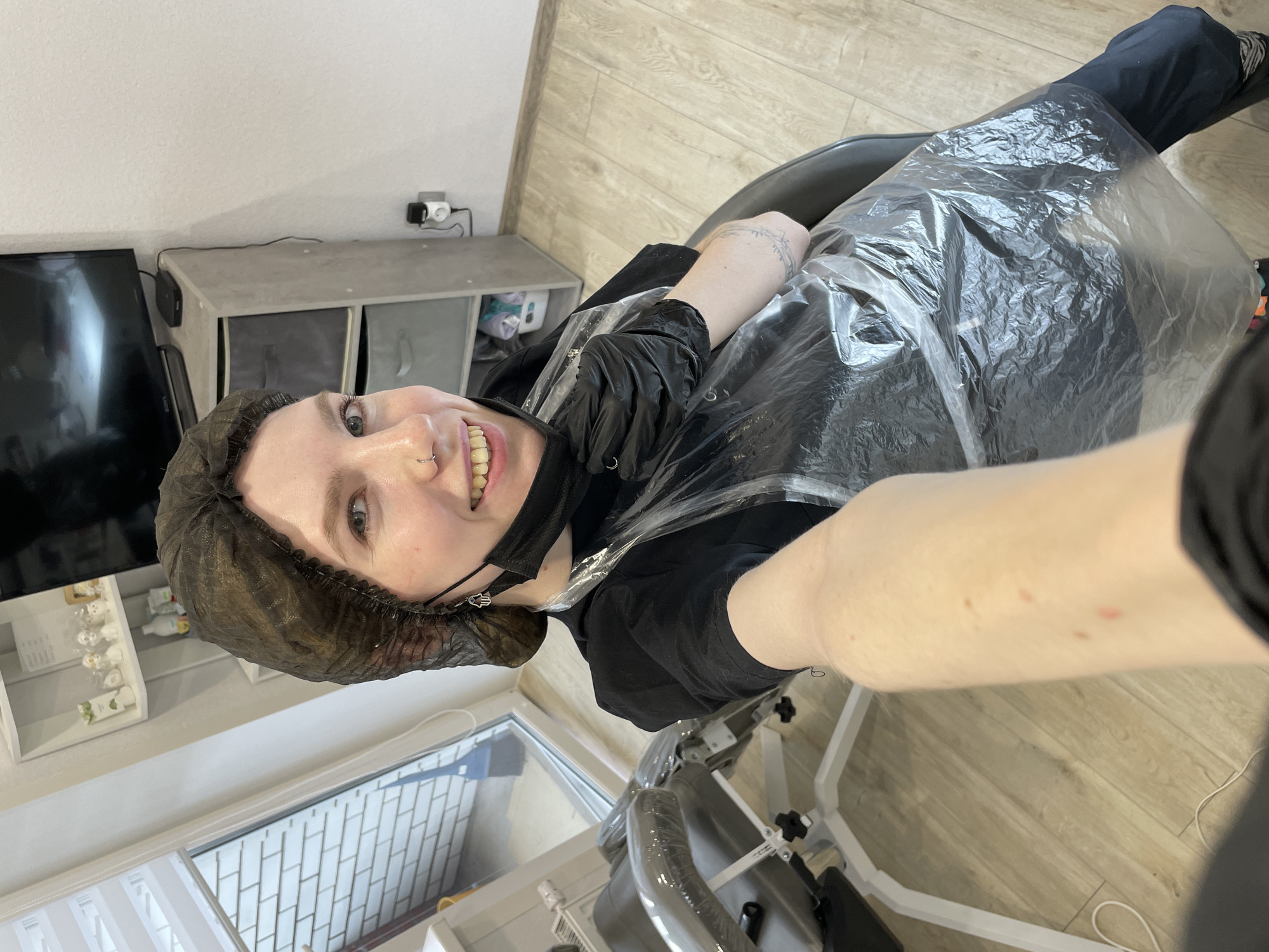 A selfie taken indoors shows a Caucasian woman with light skin, wearing a dark brown hairnet and a black face mask. She is smiling and looking up at the camera. The woman is also wearing a black t-shirt and black gloves. A transparent plastic sheet covers her body and the chair she is sitting in. The background includes a window with white blinds, a white shelf unit with bottles, a black TV, a grey cabinet with storage bins, and a light brown wooden floor. While the location isn't directly discernible from the image, the user's request indicated the city is Izhevsk, Russia.