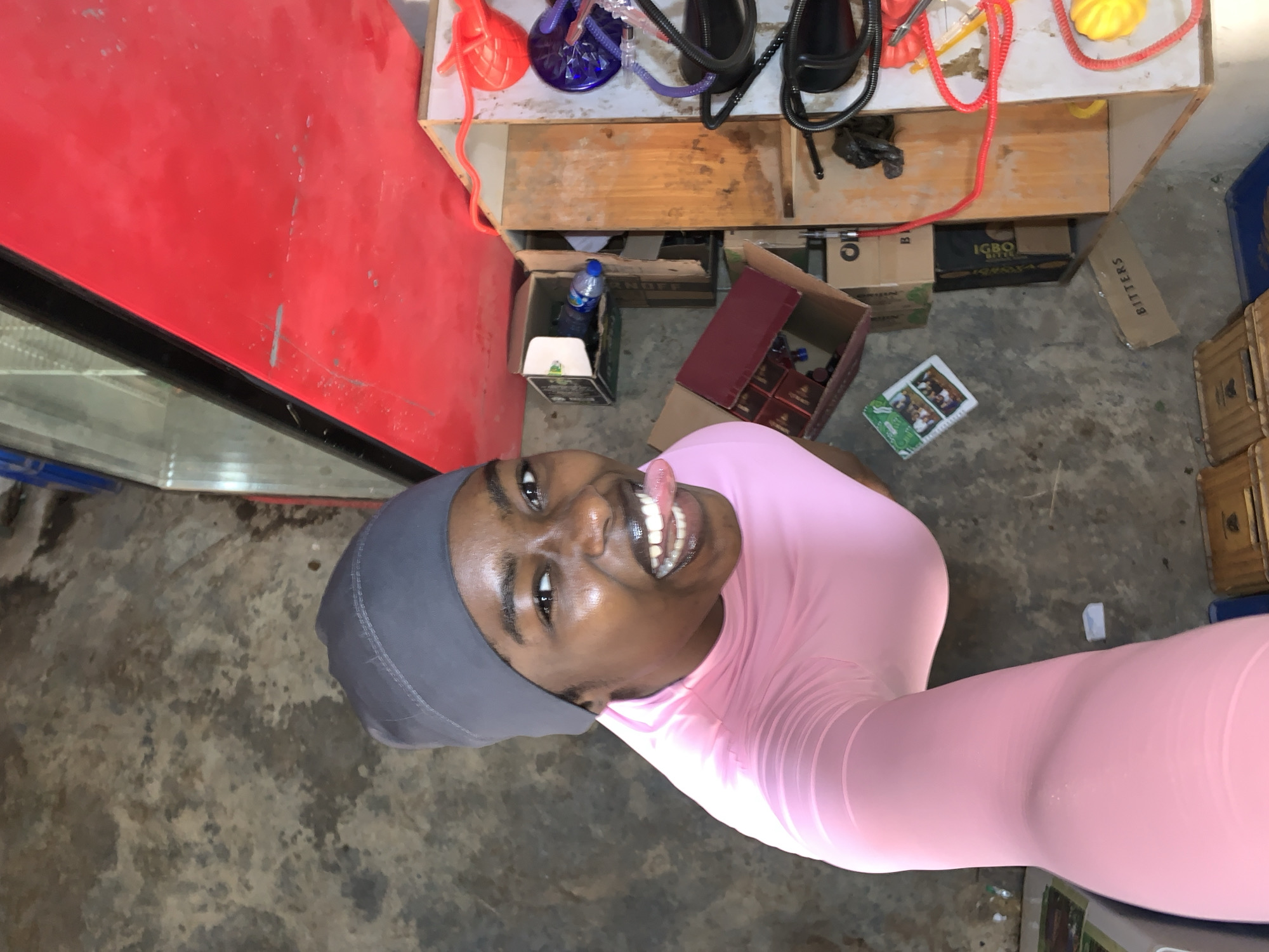 The photograph shows a person with dark skin, wearing a gray head covering and a pink long-sleeved shirt. The person is facing upward, sticking out their tongue. They are standing near a red refrigerator and a wooden shelf that contains what appear to be hookahs and their accessories. Several boxes and crates are on the floor. The location is inferred to be in New Stadium, Nigeria, based on the prompt.