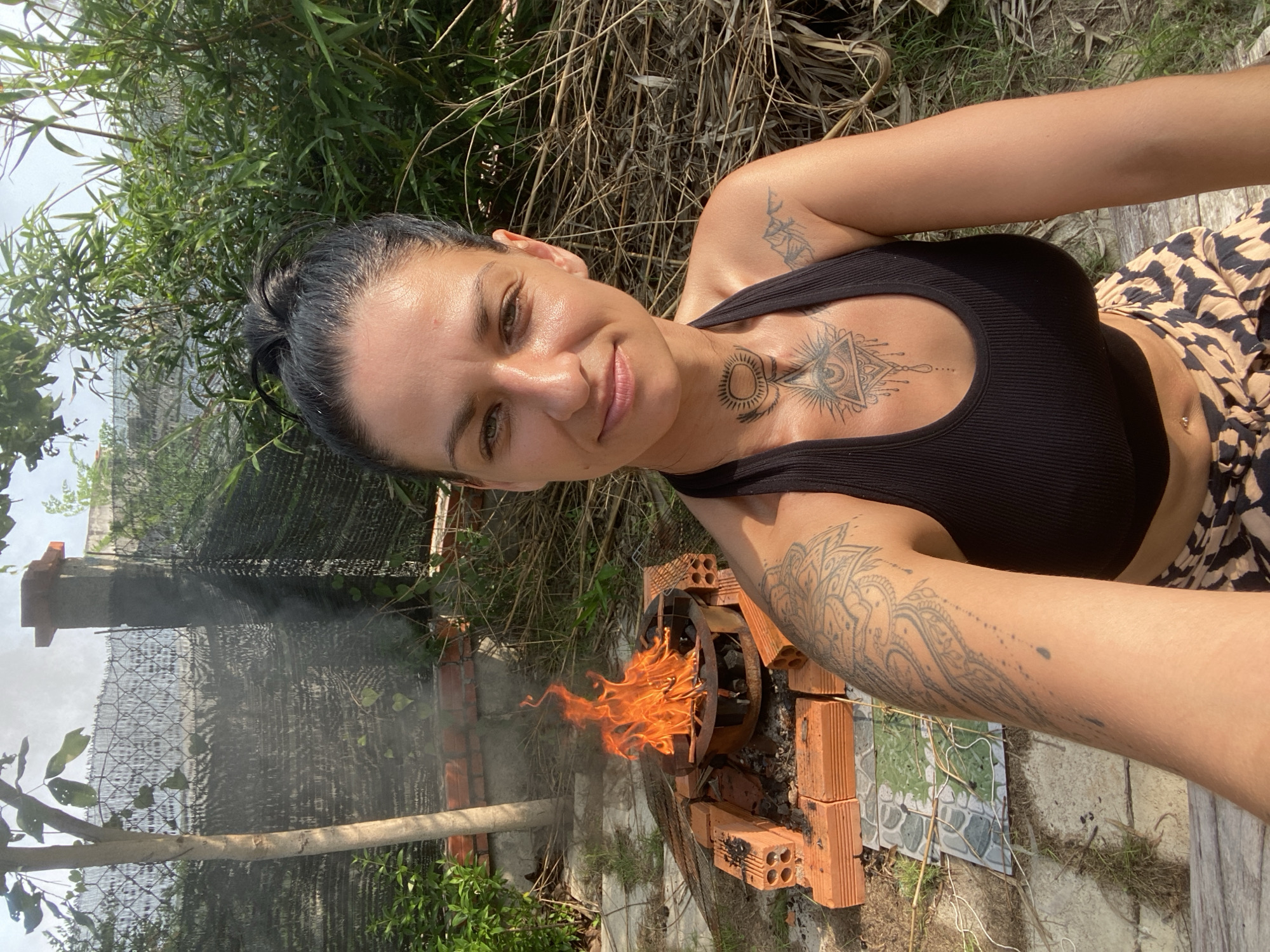 In Mũi Né, Vietnam, a woman with dark hair pulled into a bun and extensive tattoos on her arms and chest poses for a selfie. She wears a black tank top and a patterned skirt. Behind her, a small outdoor fire burns in a makeshift grill constructed from bricks and metal. The background features lush green foliage and a chain-link fence, suggesting a backyard or rural setting. Sunlight illuminates the scene.
