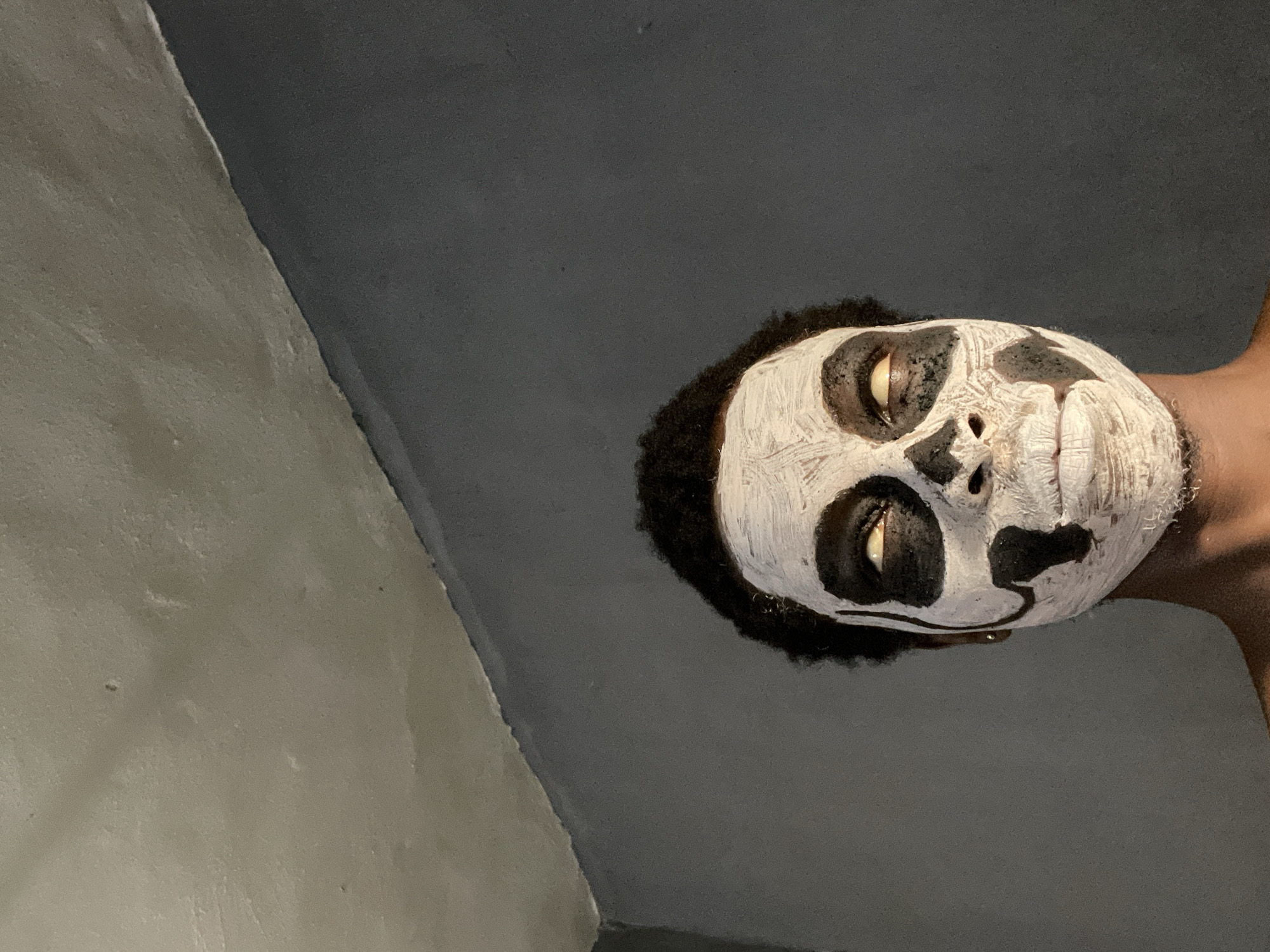 A man with black hair is looking down at the camera. His face is painted white with black circles around his eyes, black markings on his nose, and black markings around his mouth to resemble a skull. The background is a plain, dark grey wall. The location cannot be determined to be Onitsha, Nigeria from the image.