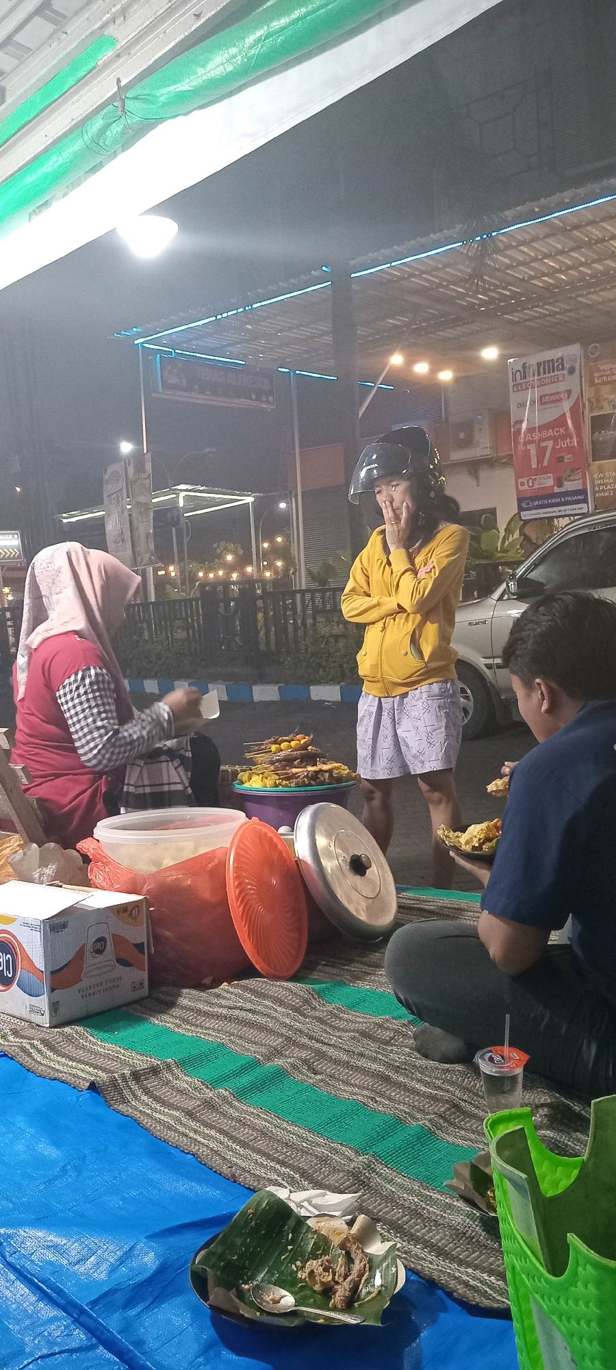 The image depicts a street food scene at night in Lamongan, Indonesia, likely near a roadside stall or outdoor dining area. In the foreground, a person in a navy blue shirt is seated on a mat, facing away from the camera, and appears to be eating from a plate. Next to them is a glass with a straw. To the left, a person wearing a pink hijab and a red and black checkered shirt is seated, holding a small white cup. A stack of satay skewers is visible on a purple tray in front of them, alongside serving bowls and containers. A young woman in a yellow hoodie and patterned shorts, wearing a black helmet, stands near the food stall, with her hand to her face. In the background, there are other stalls, a car, and decorative lighting, suggesting a lively evening atmosphere. The overall environment appears to be a casual, open-air dining setting.