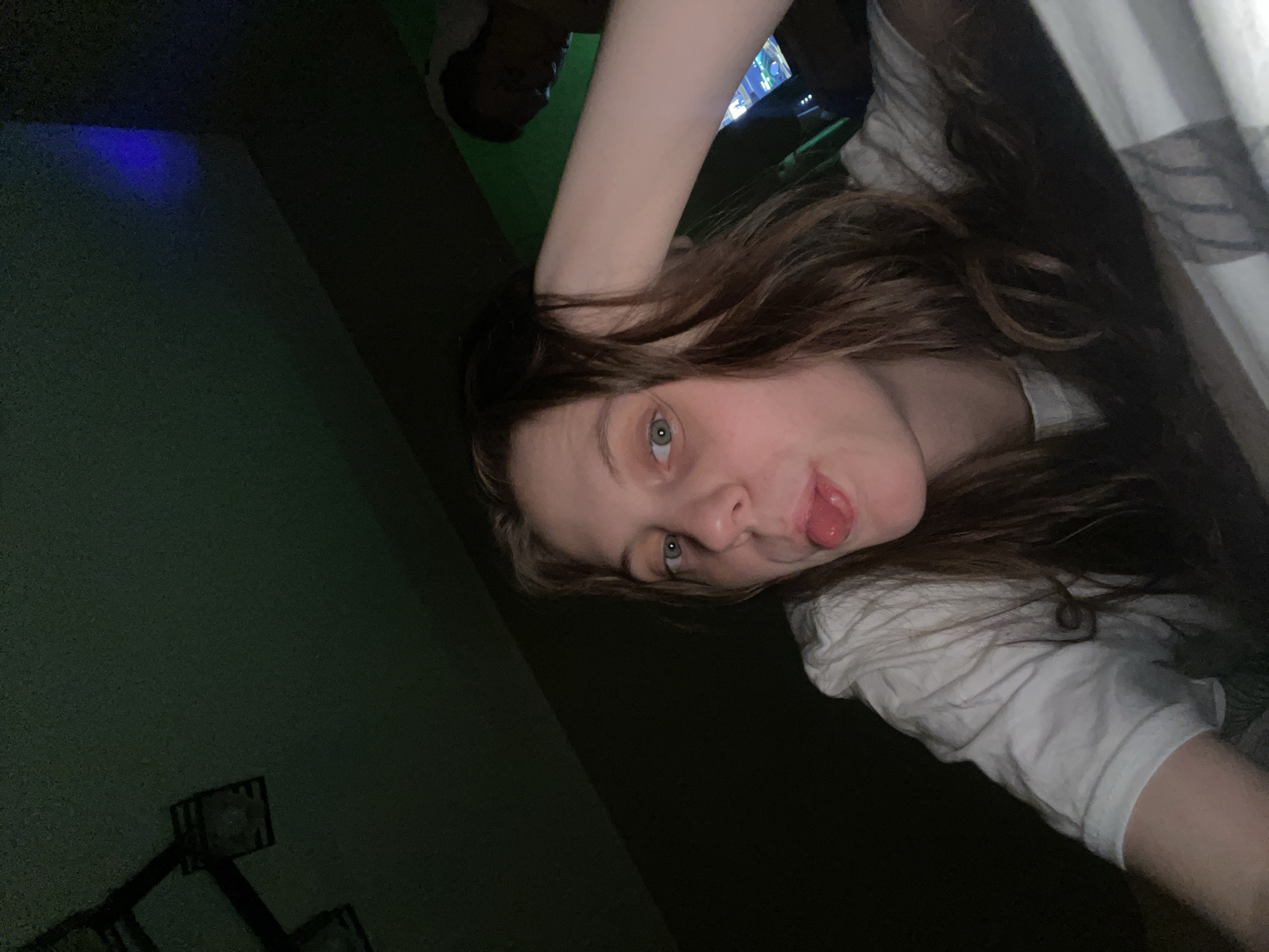 A woman is posing for a selfie indoors. She is sticking her tongue out. She has brown hair and blue eyes. She is wearing a white t-shirt. There is another person in the background. The location is not discernable from the image.