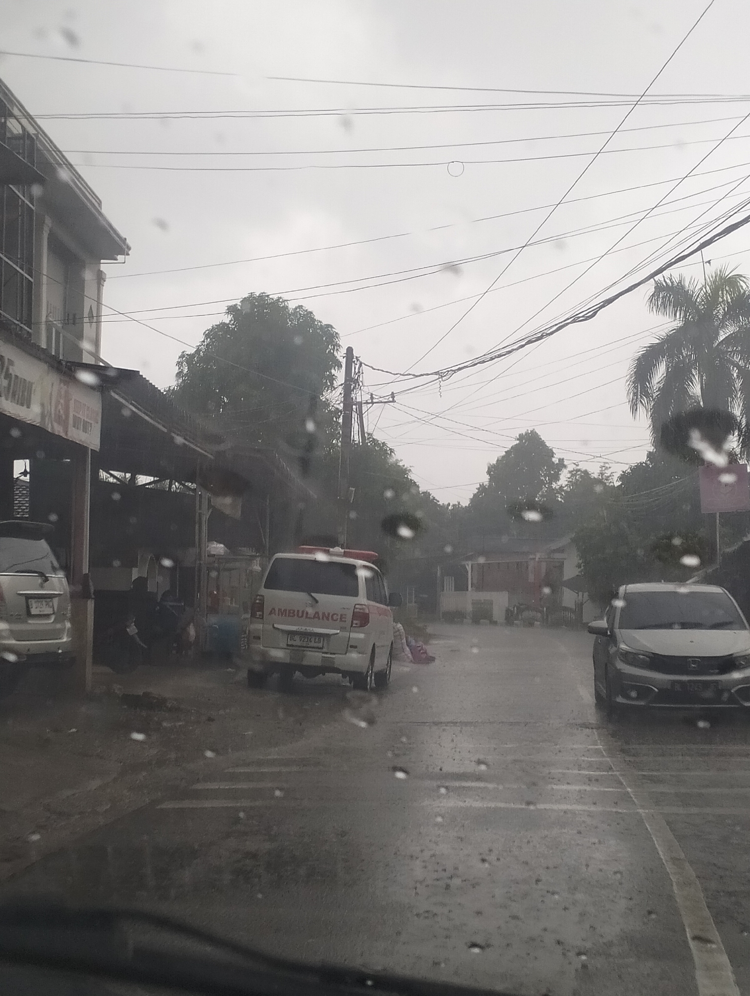 The image depicts a street scene in Banda Aceh, Indonesia, during a heavy downpour. The weather is overcast and rainy, with numerous water droplets visible on the foreground, suggesting the photo was taken from inside a vehicle.
In the foreground, the road is wet and reflects the dim light. To the left, part of a white SUV is visible. Further down the street, an ambulance with red and white markings and flashing lights is parked. The word "AMBULANCE" is clearly visible on its side. A license plate reads "BL 9234 LB". Next to the ambulance, there are some people sitting under a covered structure.
On the right side of the street, a silver car is parked. The street is lined with buildings and trees, which appear lush and green, indicating a tropical environment. Power lines are strung across the sky, a common sight in urban and semi-urban areas. The overall atmosphere is somber due to the rain and the cloudy sky. The presence of the ambulance suggests a possible emergency or a medical transport situation. The time of day is unclear due to the overcast conditions, but it appears to be daytime.