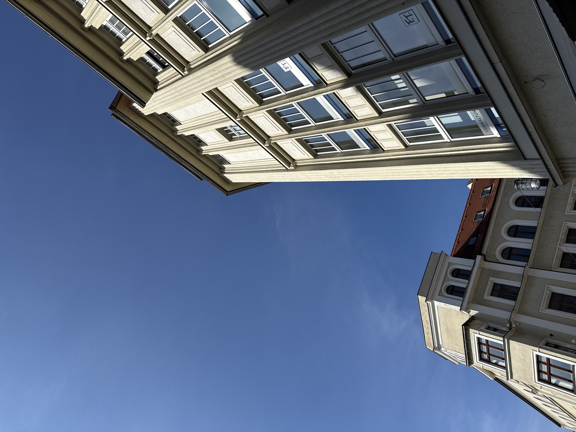 The image shows an upward angle view of two buildings against a clear blue sky. The building on the right is a modern, light-colored structure with a facade featuring prominent vertical fluting and rectangular windows with white frames. Some windows have a partial tint, and one window displays a logo with the letters "FT" within a square. Another window has the word "VERWALTUNG" printed on it. The building on the left is an older, lighter-colored building with a more ornate facade, characterized by arched windows with darker frames and decorative white trim. Its roof is reddish-brown. The setting appears to be a street in Görlitz, Germany, given the architectural style and the presence of signage in German. There are no people visible in the image. The lighting suggests it is daytime, with sunlight casting shadows on the buildings.