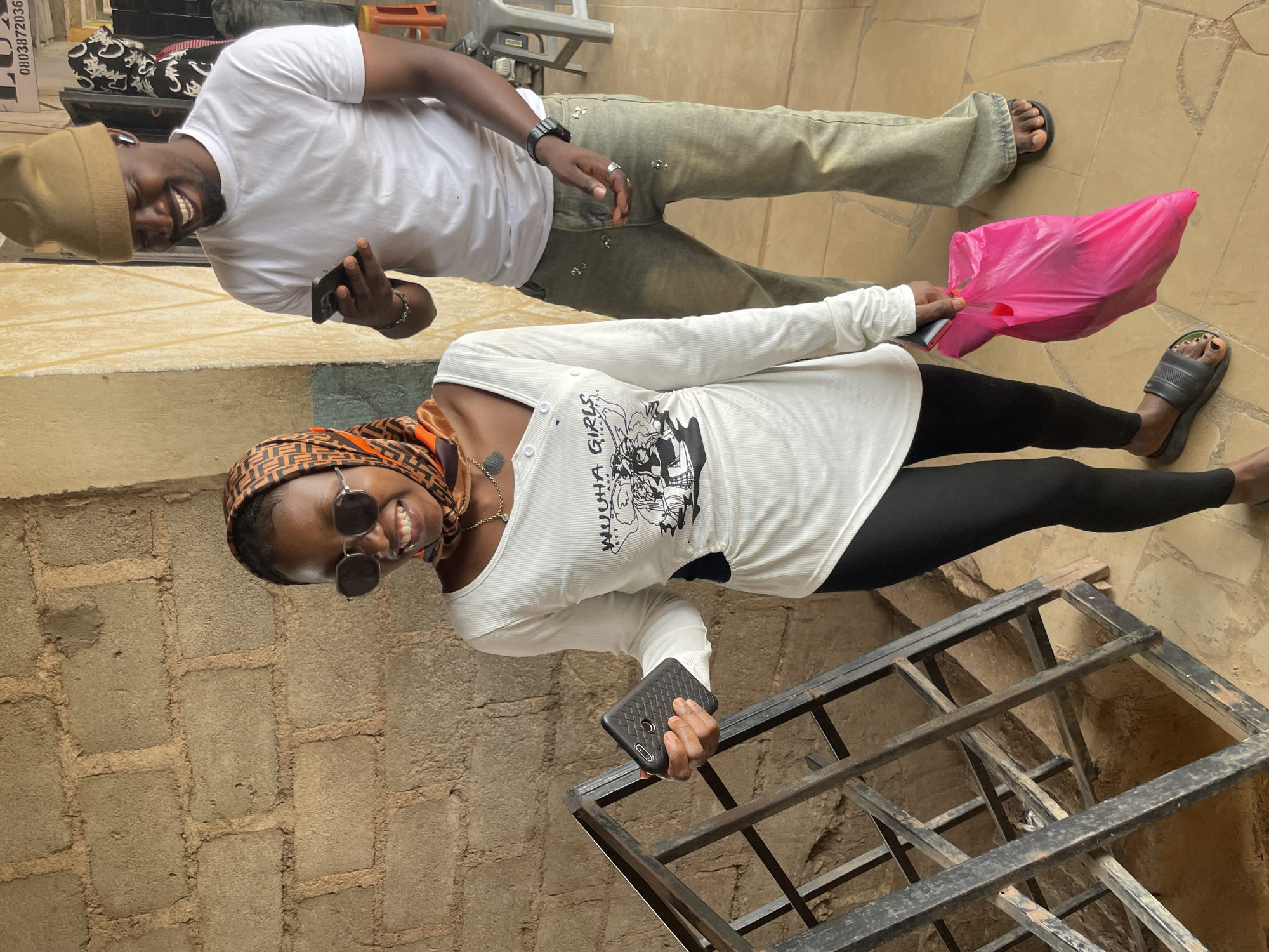 The image shows two individuals, a man and a woman, standing on a tiled patio. The man, wearing a white t-shirt, a tan beanie, and light-colored pants, is smiling and holding a smartphone. The woman, dressed in a white long-sleeved shirt and black leggings, is also smiling and holding a smartphone. She is wearing sunglasses and a patterned headscarf. A bright pink plastic bag is visible near her. The background consists of a brick wall and a metal grate. The overall impression is of a casual outdoor encounter, possibly during daylight. The image is oriented sideways.
