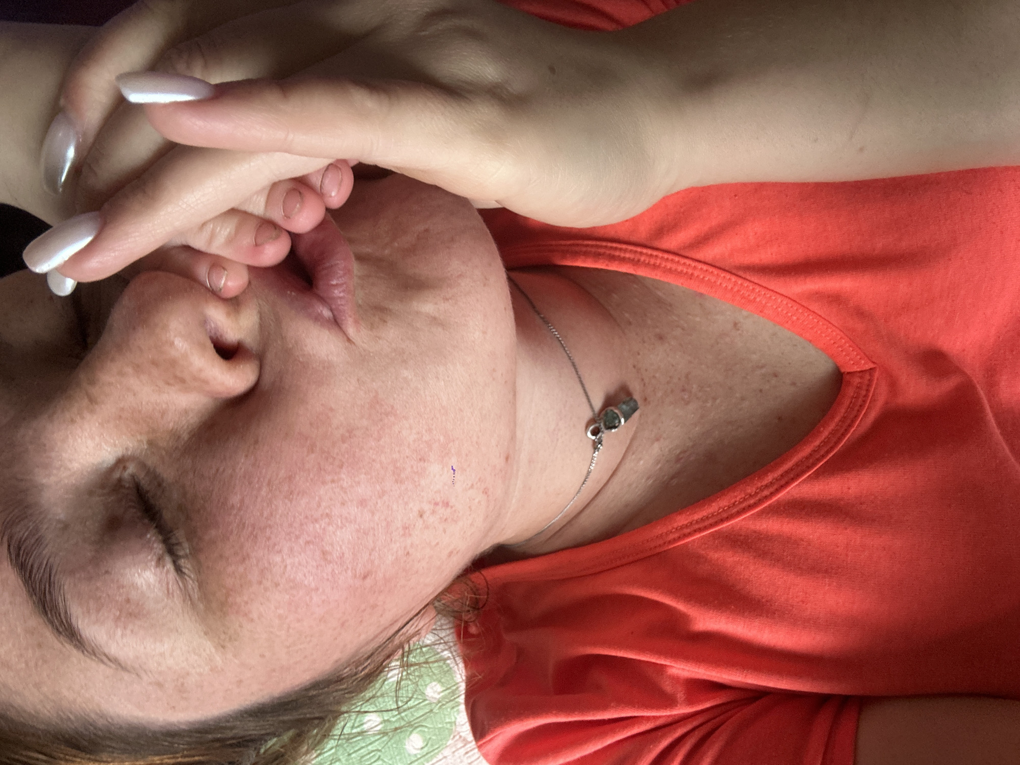 The image shows a close-up, overhead view of a person lying down, with their face partially obscured by their hands. The person has fair skin with freckles, and appears to be wearing a coral-colored tank top. A delicate silver necklace with a small pendant is visible around their neck. The person's fingernails are long, white, and manicured. The background is a light green and white patterned fabric, possibly a blanket or bedsheet. The lighting suggests an indoor setting, possibly during the daytime. The overall impression is one of rest or repose, with the hands covering the face perhaps indicating a moment of introspection or sleep. There is no visible text in the image. The location context is Nizhny Novgorod, Russia, though no specific landmarks or cues confirm this within the image itself.