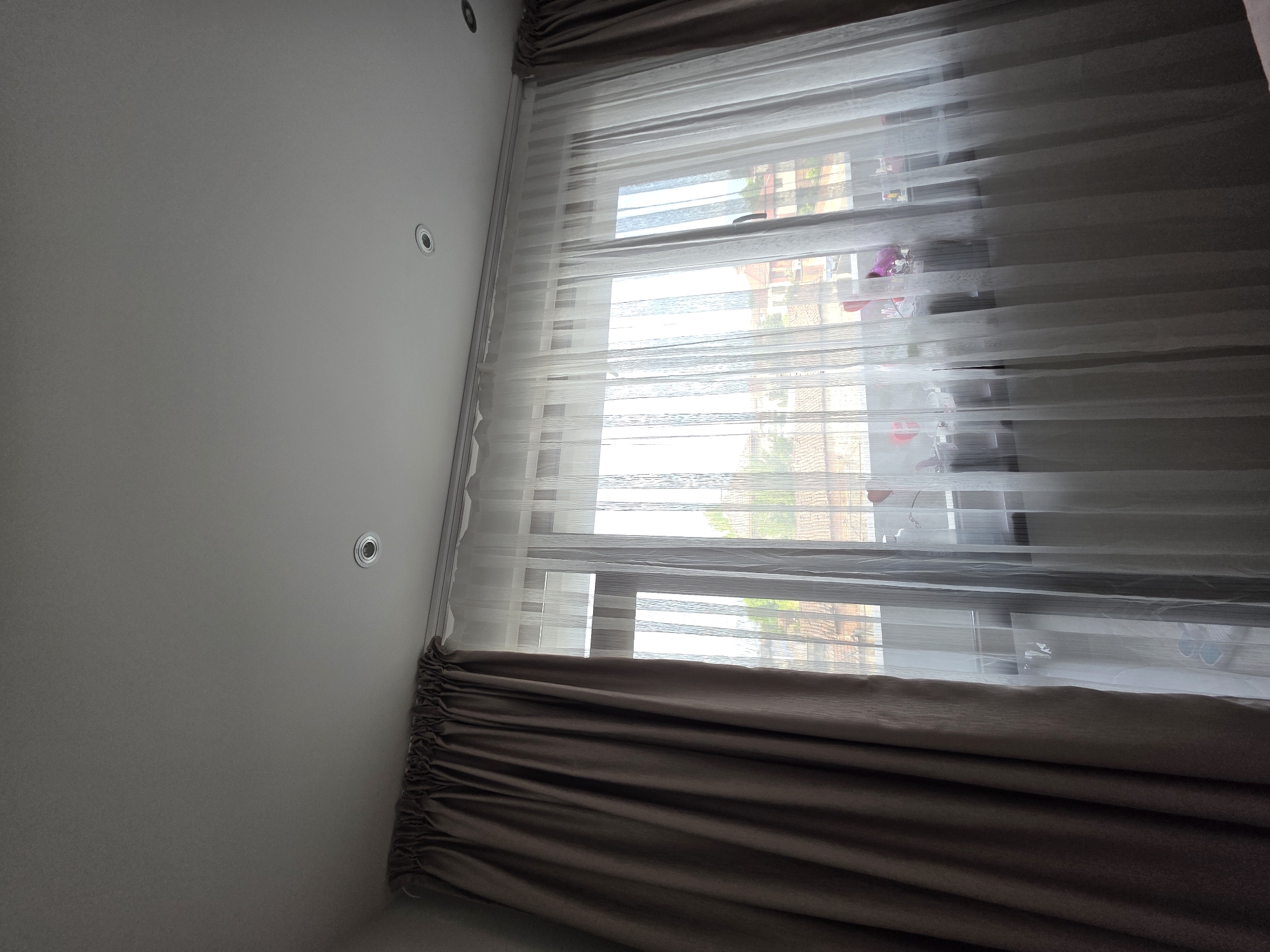 The image shows a window with layered curtains in an interior setting. The outermost layer consists of heavier, brown drapes that are partially drawn. A sheer, white curtain is positioned behind the brown drapes. The window reveals a view of the outside, possibly a residential area, with buildings visible through the semi-transparent curtains. The ceiling is visible, featuring recessed lighting fixtures. The location is stated as Yagodovo, Bulgaria.