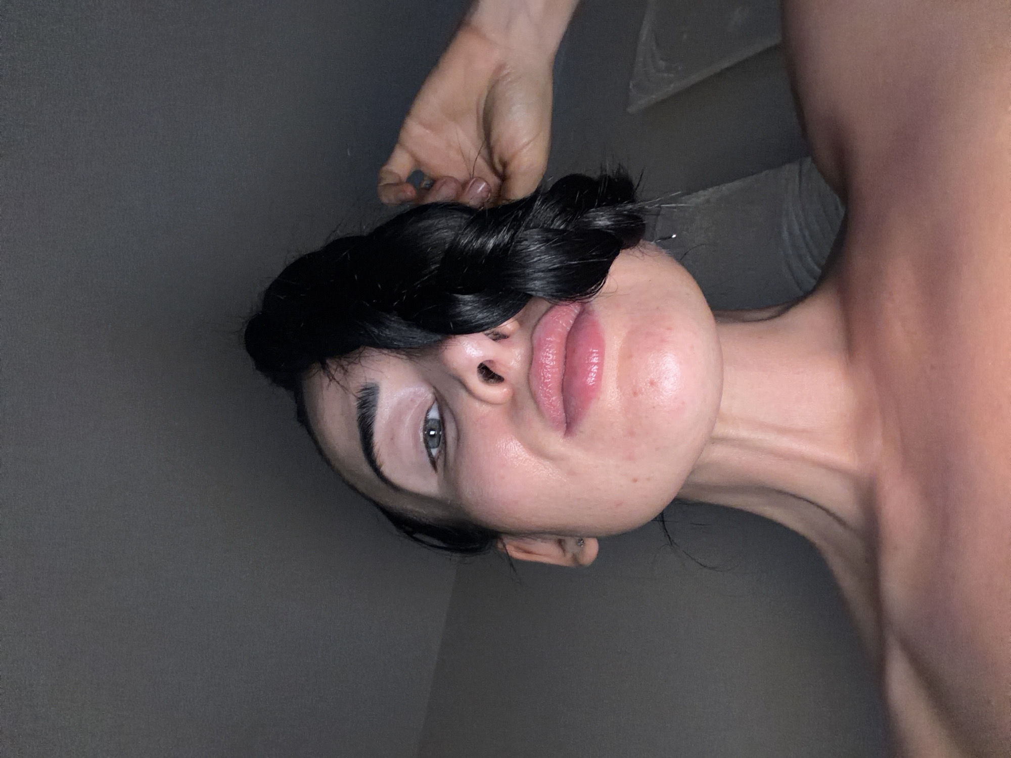 A close-up, vertically oriented portrait features a young woman with dark hair. Her head is angled, and she appears to be looking upwards and slightly to the left. Her dark hair is styled, possibly in a thick braid that curves around the side of her head. Her left eye, from the viewer's perspective, is visible and appears to be a light blue or grey color, with dark eyebrows framing it. Her full lips are slightly parted, showcasing a glossy pink hue.
Her skin has a natural appearance, with visible pores and some minor redness or blemishes on her cheek and chin. A hand with short fingernails, one of which appears to have dark polish, is positioned above her head, gently touching her hair. Her neck and a bare shoulder are visible on the right side of the frame, suggesting she is either unclothed or wearing something that exposes her upper body.
The background is a plain, dark grey surface, offering no specific location details beyond being indoors. The lighting is soft and somewhat diffuse, highlighting the contours of her face and neck. The overall impression is one of calm and introspection. No text is visible in the image.