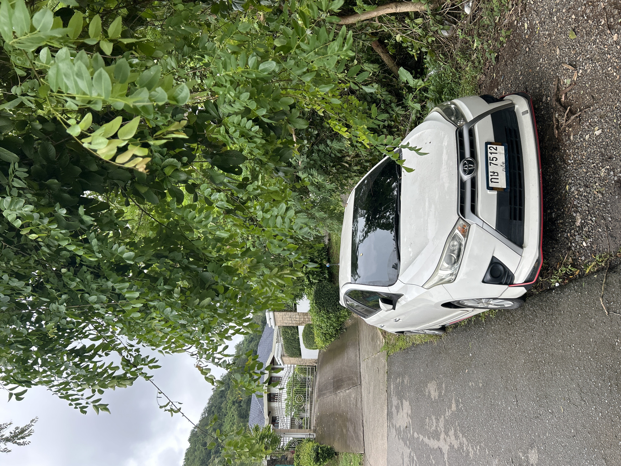 A white Toyota Yaris is parked on the side of a wet, asphalt road in Kathu, Thailand. The car is facing towards a large, white house with a dark tiled roof, accessible via a concrete driveway with an ornate metal gate. The scene is heavily shaded by lush green trees and foliage, with a hilly, vegetated landscape visible in the background. The sky is overcast. The car's license plate reads "กข 7512".