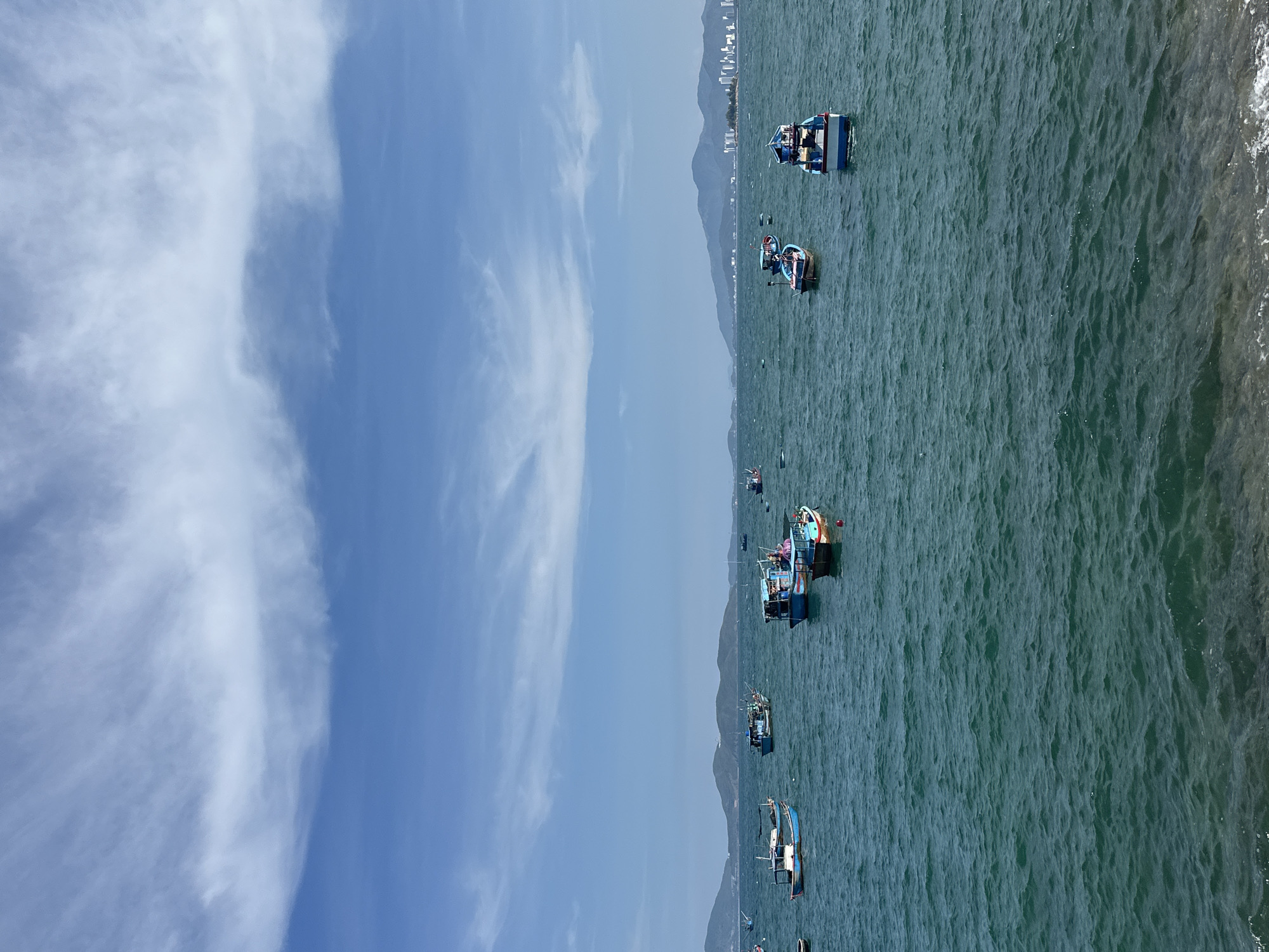 The image depicts a coastal scene in Bac Nha Trang Ward, Vietnam, featuring a body of water with several small fishing boats scattered across the surface. The water appears choppy with discernible waves. In the background, the coastline is visible, characterized by a mountain range and cityscape. The sky is partially covered with light clouds against a blue expanse. No people are clearly visible on the boats.