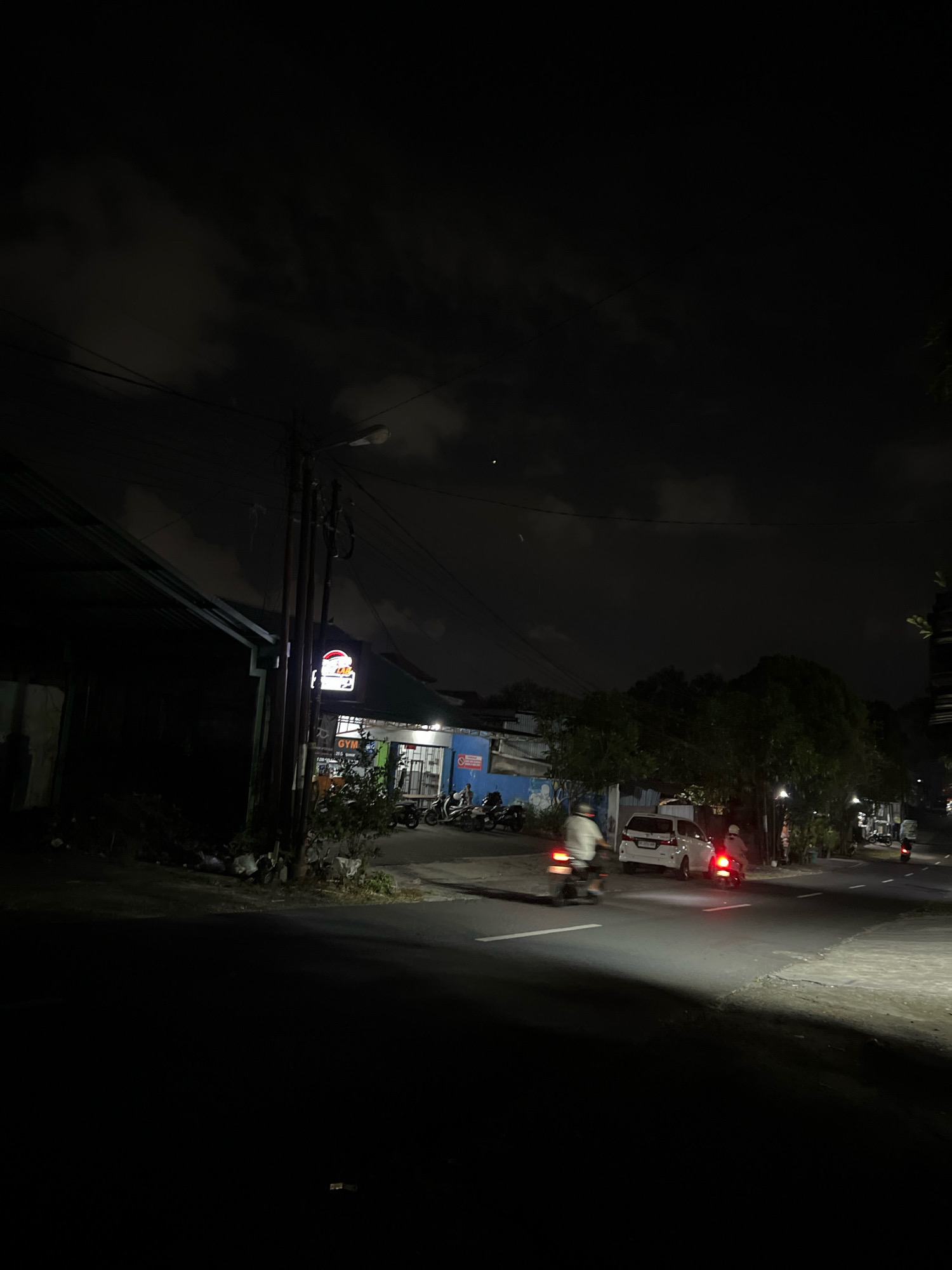 The image depicts a nighttime street scene in Denpasar, Indonesia, likely in a commercial or residential area. A dark, overcast sky looms above, with visible clouds illuminated by ambient light. A street lamp on a utility pole casts a downward glow.
In the foreground, a paved road is visible, with its surface reflecting the headlights of a white vehicle parked on the right side of the road. The vehicle's brake lights are illuminated red. Two individuals are seen riding motorcycles on the road, their forms blurred by motion. One motorcycle is in the left lane, moving away from the viewer, while another is in the right lane, also moving away from the viewer, its brake lights brightly lit.
Along the left side of the road, a dark, low-lying structure with a corrugated metal roof is partially visible. Adjacent to this structure, a building with a blue facade is present, displaying a lit sign with red and white lettering, possibly indicating a business or establishment. Several parked motorcycles are visible in front of this building. Trees and vegetation line the right side of the road, obscuring the background. The overall impression is of a typical urban street at night, with ongoing activity despite the darkness.