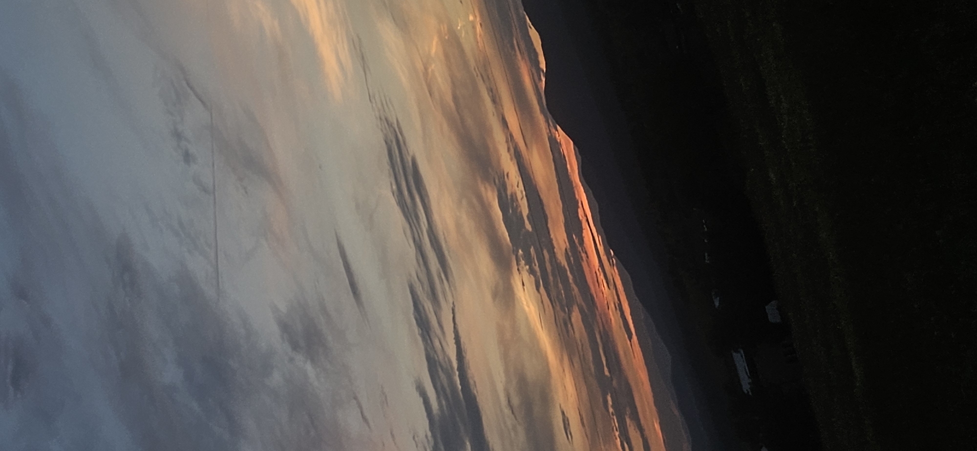 The image depicts a dusky sky over rolling hills, with the sun having recently set. The sky is a gradient of blues, grays, and oranges, with wispy clouds streaking horizontally. In the foreground, dark silhouettes of mountains recede into the distance. The lower portion of the image shows a dark, grassy landscape with faint outlines of buildings or structures, possibly a small village or rural settlement. The scene conveys a sense of natural beauty and tranquility, characteristic of a rural or mountainous region at dusk. The specific location in Momin Prohod, Bulgaria, is not discernible from the visual elements of the image alone.