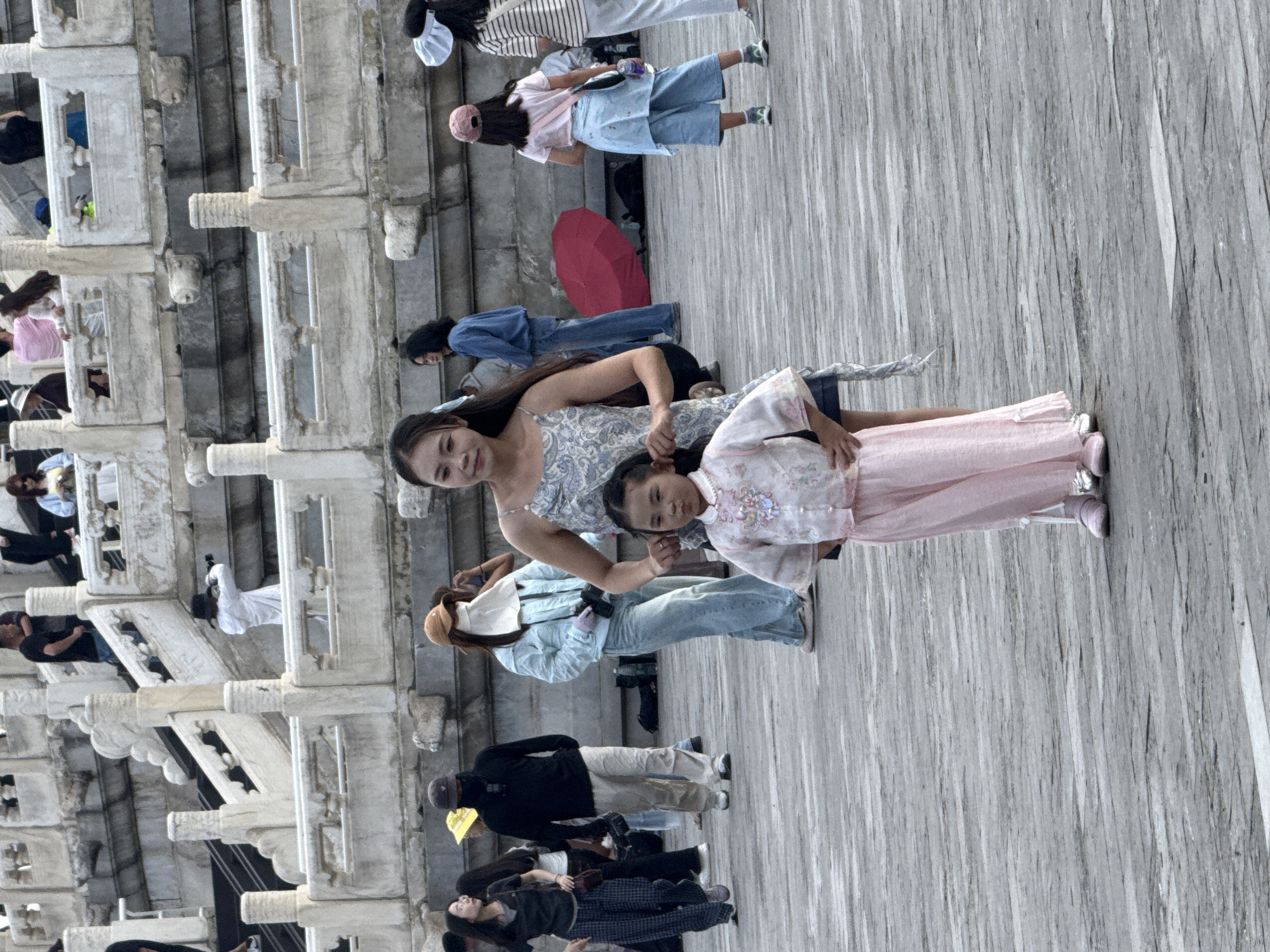 In Dongcheng District, China, at an outdoor location with marble architecture, a woman in a patterned dress stands with a young girl in a traditional pink outfit. The woman has her hand on the girl's back, while the girl stands with her hands on her hips. Several other individuals are visible in the background, some walking, some standing, and one taking a photo. A red umbrella lies on the ground near a person wearing a light blue jacket and jeans, who is holding a camera. The ground is paved with wooden planks, and the architectural elements suggest a historical or monumental site.