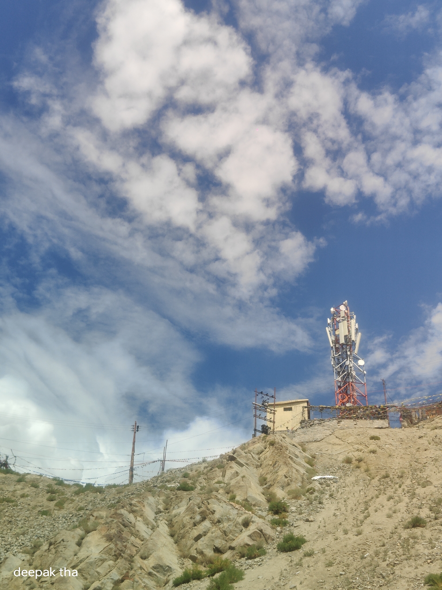 The image depicts a cellular tower situated on a rocky, barren hillside. The tower is tall and multi-faceted, with various antennas and communication equipment attached to its structure. Below the tower, there's a small, light-colored building. The hillside is arid, with sparse vegetation and scattered rocks. Prayer flags are strung along a line on the hillside, suggesting a cultural or religious significance to the location. The sky above is a vibrant blue, with scattered white clouds. The watermark "deepak tha" is visible in the lower left corner of the image. This scene is characteristic of the high-altitude regions surrounding Leh, India, where such communication infrastructure is often built on elevated terrain.
