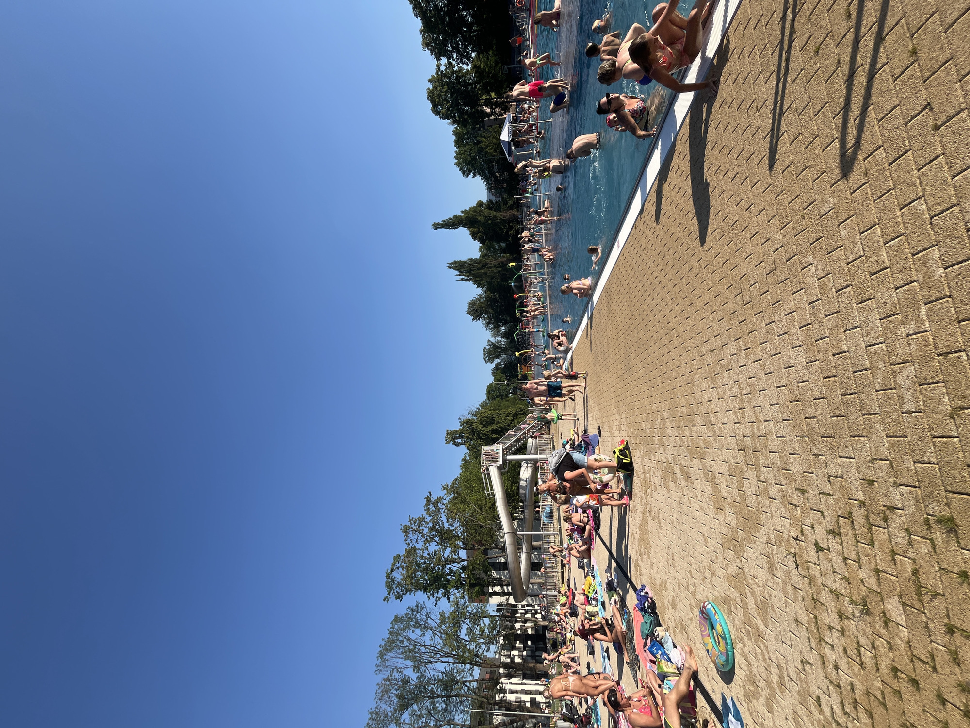 This media file depicts a lively outdoor swimming pool scene on a sunny day, indicated by the clear blue sky and the strong shadows cast on the brick-patterned ground. The image is oriented vertically, with the pool occupying the left half and the sunbathing area on the right.
Numerous people are enjoying the pool, some are swimming, others are sitting or standing at the edge. A water slide structure is visible in the background, with people interacting with it. The sunbathing area is populated with many individuals relaxing on towels and blankets, soaking up the sun. Various beach items such as towels, bags, and a colorful inflatable ring are scattered around.
The overall atmosphere appears to be one of leisure and enjoyment, with a strong sense of summer vacation or a recreational outing. The weather is clearly warm and sunny, suggesting midday or afternoon. The abundance of people and their relaxed postures convey a sense of a popular public space during peak season. There is no visible text in the image.