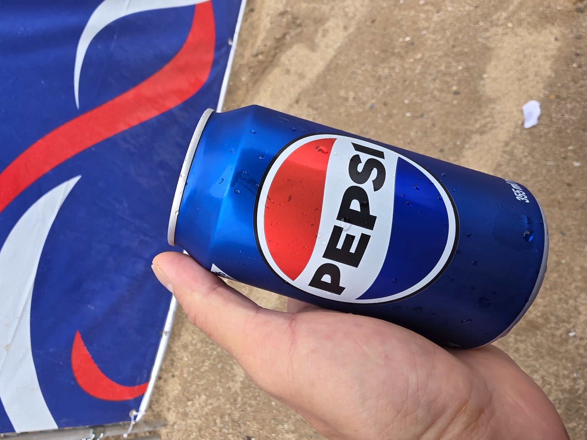 A person's hand is holding a cold, condensation-covered blue Pepsi can. The can has the iconic Pepsi logo, featuring red, white, and blue circles, with the word "PEPSI" prominently displayed in white letters. In the background, a blue banner with red and white wave-like designs is visible. The ground appears to be sandy or dirt-like. The scene is outdoors, likely in an urban environment given the presence of a banner, suggesting a public event or advertisement. There is no specific indication of the location within Daejeon, South Korea, beyond it being an outdoor space.