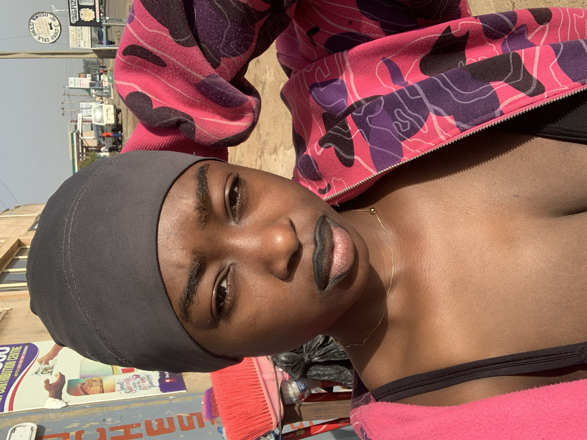 A close-up, angled shot of a dark-skinned woman wearing a dark gray head covering and a pink and purple camouflage-patterned jacket. She has dark lipstick and a delicate gold chain around her neck. The background shows an urban environment, including a sign that reads "Small Savings Dreams Tomorrow," advertising a contribution center. A sign above it is partly obscured but has the words "CHIEF" and "UNISEX" visible. The presence of street signs and a vehicle in the background suggests a city street. Jos, Nigeria is written on a sign in the background. A red broom is in the foreground on the bottom left.