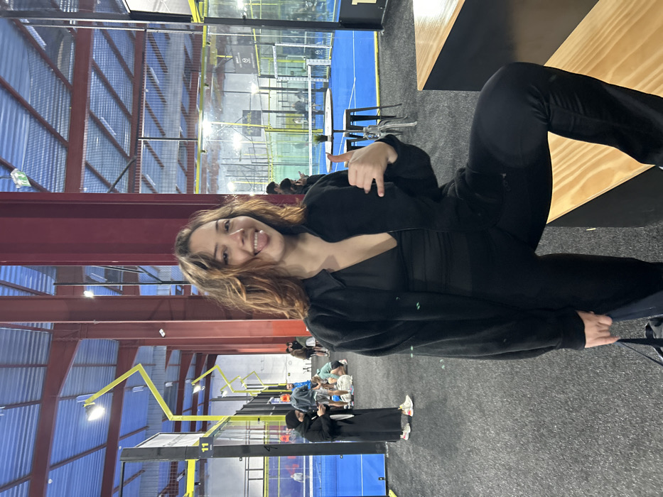 The image displays a young woman smiling and posing for the camera inside an indoor padel court facility in Eckbolsheim, France. She has light brown, wavy hair and is looking directly at the viewer with a happy expression, revealing a tongue piercing. She is wearing a black zip-up jacket over a black top and black pants, and makes a "shaka" or "hang loose" hand gesture with her right hand.
In the background, several blue padel courts with glass walls and nets are visible. Multiple other individuals are scattered throughout the facility, some seated at tables or benches, while others stand near the courts, suggesting they are either playing, spectating, or resting. One person near the lower-left is dressed in a long, dark garment.
The setting is an indoor sports complex characterized by a high ceiling with exposed red and yellow metal support beams and bright overhead lighting. Wooden tables or benches are present in the foreground and midground. A white number "11" is visible on a black sign or divider in the lower-left, and the word "PADEL" can be faintly seen on a black structure behind the main subject, adjacent to one of the courts. The flooring is a dark, textured material. The overall scene suggests a casual and active environment within a modern sports facility.