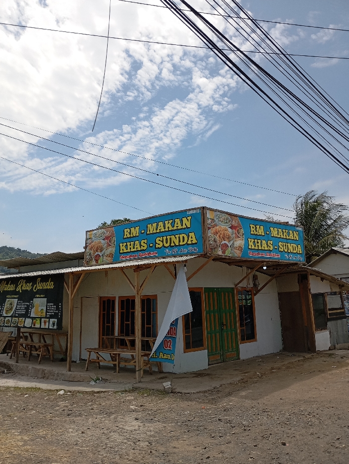 A small restaurant building, with a blue and white sign reading "RM-MAKAN KHAS-SUNDA" in Indonesian, located in Kecamatan Kutawaringin, Indonesia. The restaurant is made of wood and has a white awning covering the front. There are three wooden tables and chairs on the ground in front of the building. A white banner is hanging on the wall. The building is located on a dirt road. There are numerous power lines stretching overhead. There is a bright, clear sky with clouds. The restaurant is situated on a small hill and there are trees in the background. A second sign is visible on the right side of the building, also reading "RM-MAKAN KHAS-SUNDA". The restaurant appears to be closed.