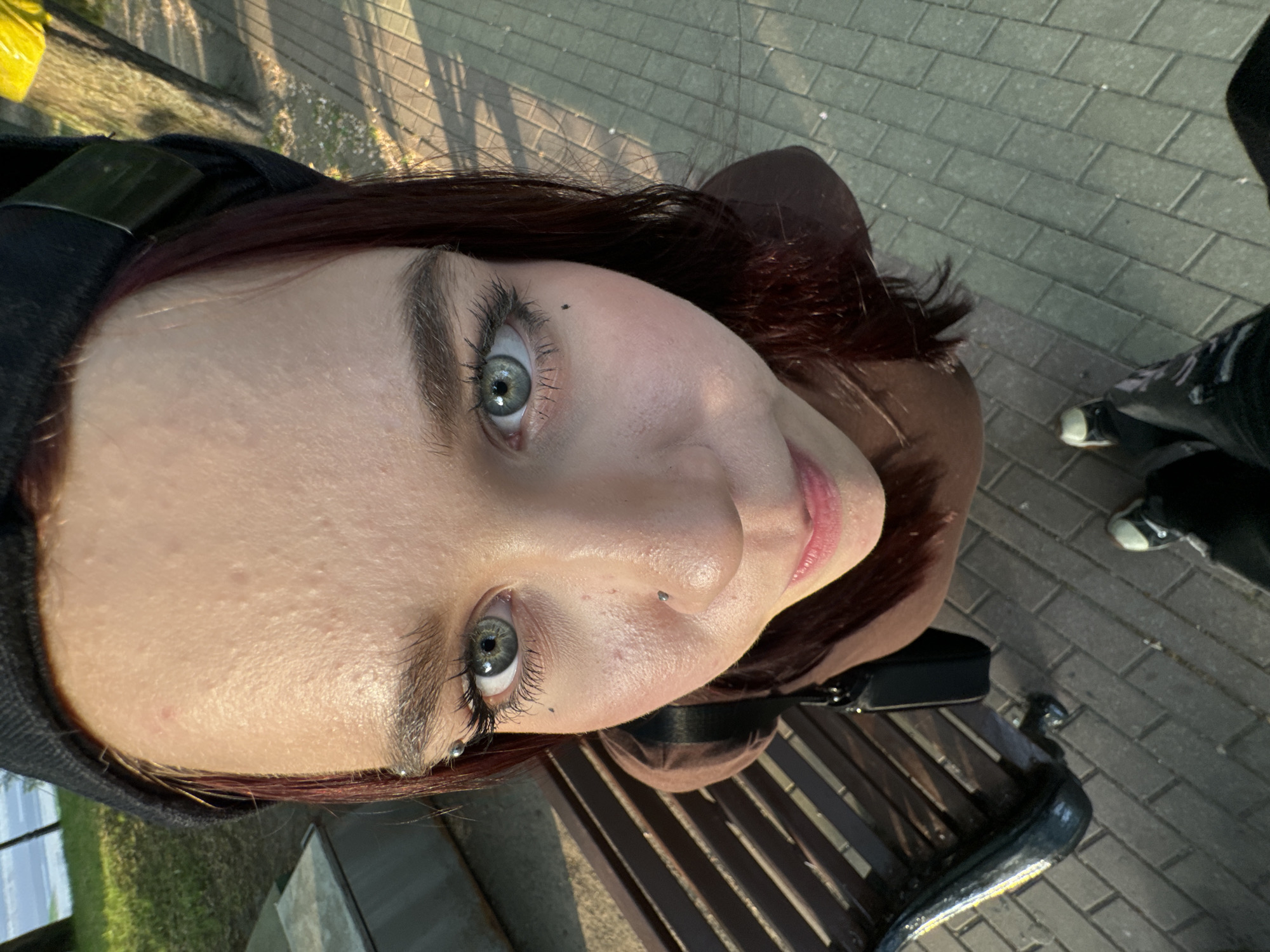 A young woman with red-tinted brown hair is captured in an upward-angle selfie. She has fair skin, gray-green eyes, and a nose piercing. Her right eye has what appears to be a dark beauty mark above the eyebrow. She is wearing a dark hooded garment over a brown hooded jacket. The woman is seated on a brown wooden park bench, and a black purse is visible beside her. In the background, there is a paved brick pathway and a glimpse of someone's black shoes and legs. The location appears to be a public area, such as a city park.