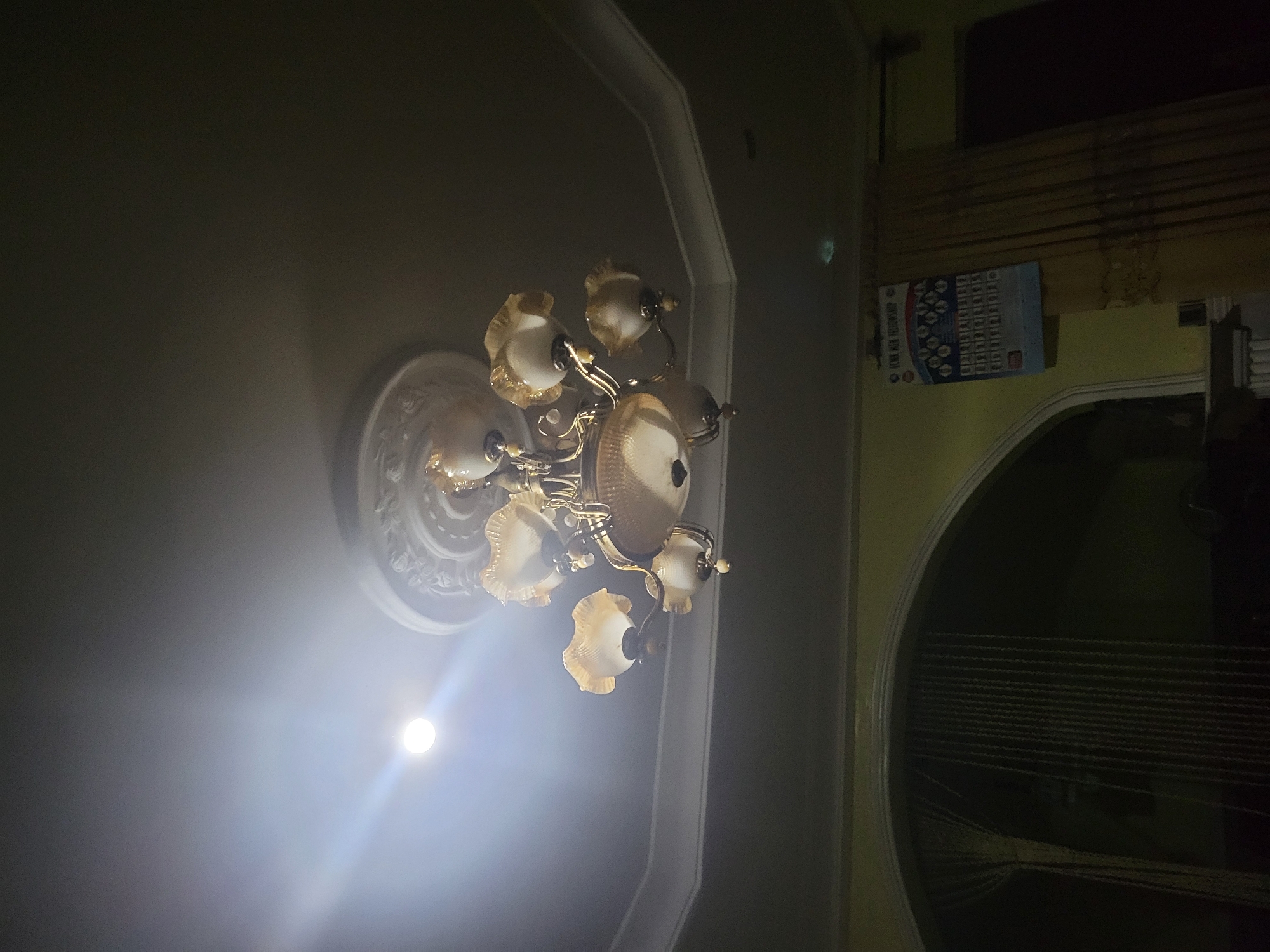 The image shows a dimly lit interior in Madalla, Nigeria. A decorative chandelier with multiple lights is affixed to a ceiling with decorative molding. A calendar is mounted on a wall. An arched doorway leads to another space with draped fabric or curtains.