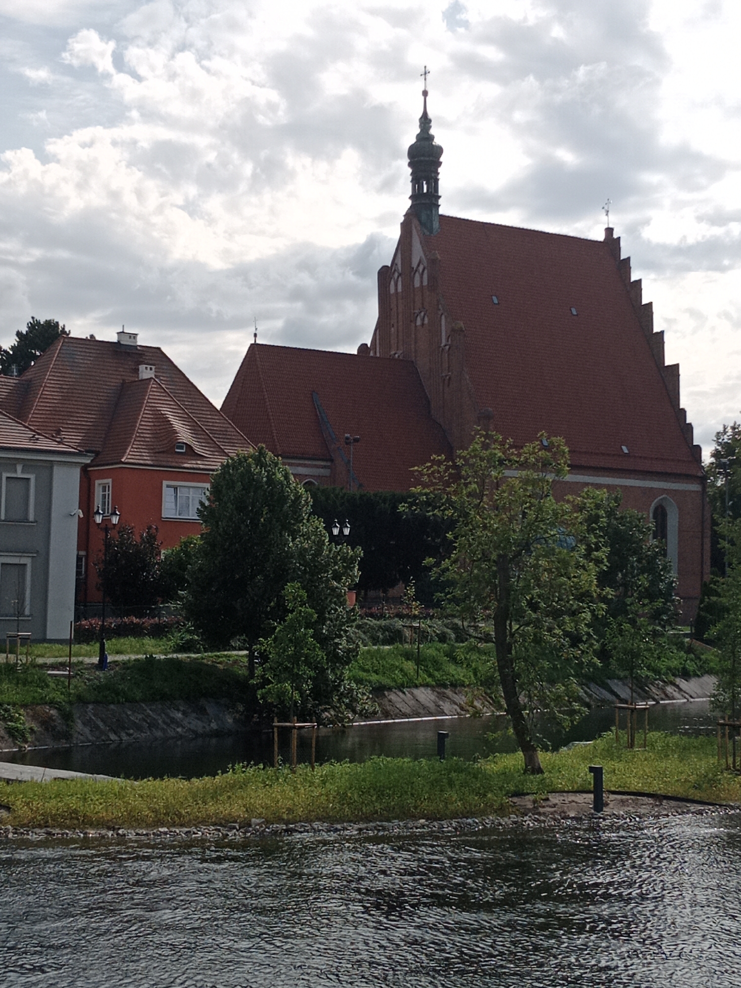 The image shows a view of the Brda River in Bydgoszcz, Poland, with a brick church and other buildings on the far bank. The church has a red tile roof and a tall steeple with a cross. There are trees and greenery along the riverbank, and the water is dark and rippling. The sky is cloudy.