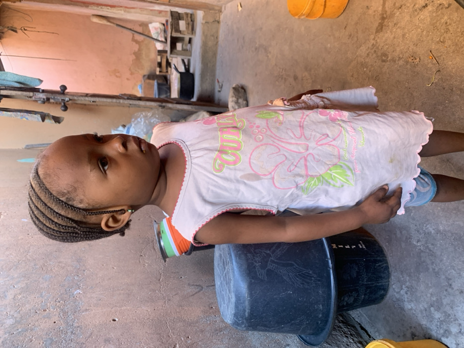 A young Black child, approximately 2-4 years old, stands facing slightly right of the camera, looking straight ahead with a neutral expression. The child has cornrow braids pulled back from the forehead and wears a light-colored, sleeveless dress with pink scalloped trim and a graphic featuring a pink hibiscus flower and green leaves. Light-blue patterned shorts are visible beneath the dress. A small green earring is present in the left earlobe.
The scene is an outdoor or semi-enclosed urban space, likely a courtyard or alleyway in Okene, Nigeria. The immediate background features a rough, textured concrete or stucco wall, painted an aged beige/light brown, exhibiting patches of wear and discoloration. To the child's left, two dark plastic buckets are stacked, the upper one inverted. Behind the child's right shoulder, a partial view of an open doorway or archway reveals a brighter, reddish-orange painted wall beyond, suggesting an interior or connecting space. Various utilitarian objects are visible in the background through this opening, including what appears to be a black cooking pot, small wooden stools or pallets, and hanging fabric. The ground is a flat, grey concrete surface with minor debris. To the far right, a stack of two yellow plastic buckets is partially visible.