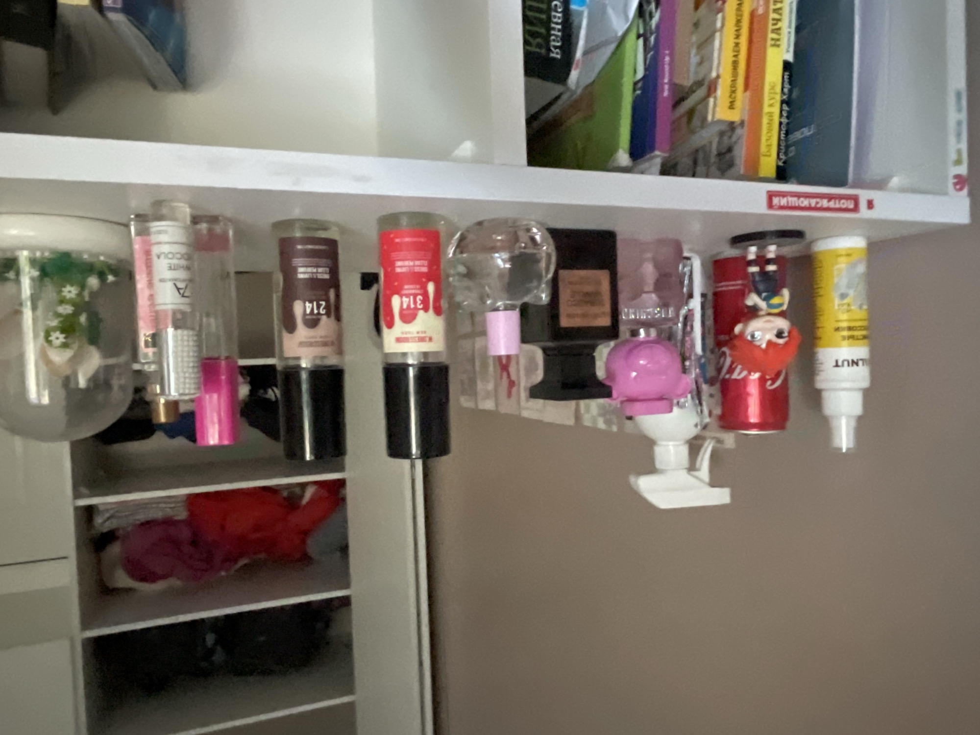 The image depicts a shelf in what appears to be a bedroom or dressing area, containing various personal care products and decorative items. On the top shelf, a white spray bottle, a red can of Coca-Cola, a pink perfume bottle shaped like a piggy bank, a black perfume bottle, and several other perfume bottles are visible. A small figurine of an anime character is also present. The shelf below contains books and binders. The overall environment suggests a domestic setting. No specific location in Мухавецкі сельскі Савет, Belarus, can be determined from the image alone.