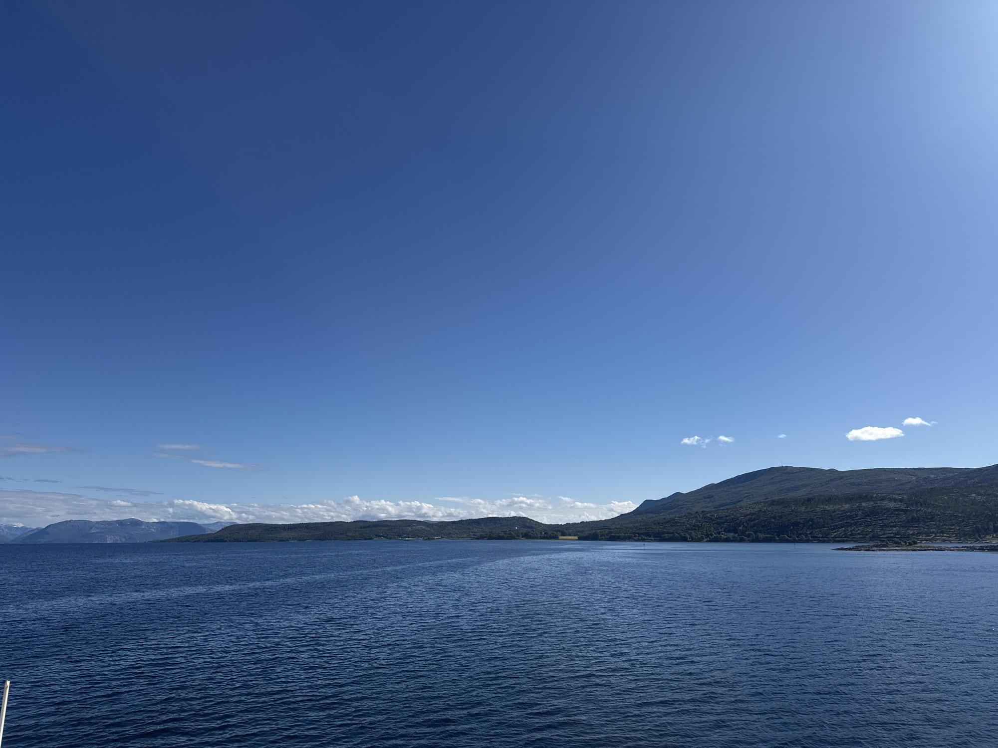 This media file is a landscape photograph taken from a boat on a body of water, likely a fjord, in Hierenjárgga - Korsnes, Norway. The scene is dominated by the deep blue expanse of the water, which has a slightly rippled surface, reflecting the clear blue sky above. In the distance, a lush, green, and hilly coastline stretches across the frame. The land is densely covered with trees and shrubs, with some rocky outcrops visible along the water's edge. The terrain rises into rolling hills and mountains, some of which are partially obscured by clouds. The sky is a vivid, cloudless blue for the most part, with a few wispy white clouds scattered near the horizon. The bright sunlight suggests it is daytime, likely mid-morning or afternoon, and the weather appears to be clear and fair. There is a sense of tranquility and natural beauty conveyed by the serene water and the majestic, tree-covered landscape. The image is composed to emphasize the vastness of the water and sky, with the coastline serving as a scenic backdrop. The overall impression is one of a peaceful day in a scenic, natural environment.