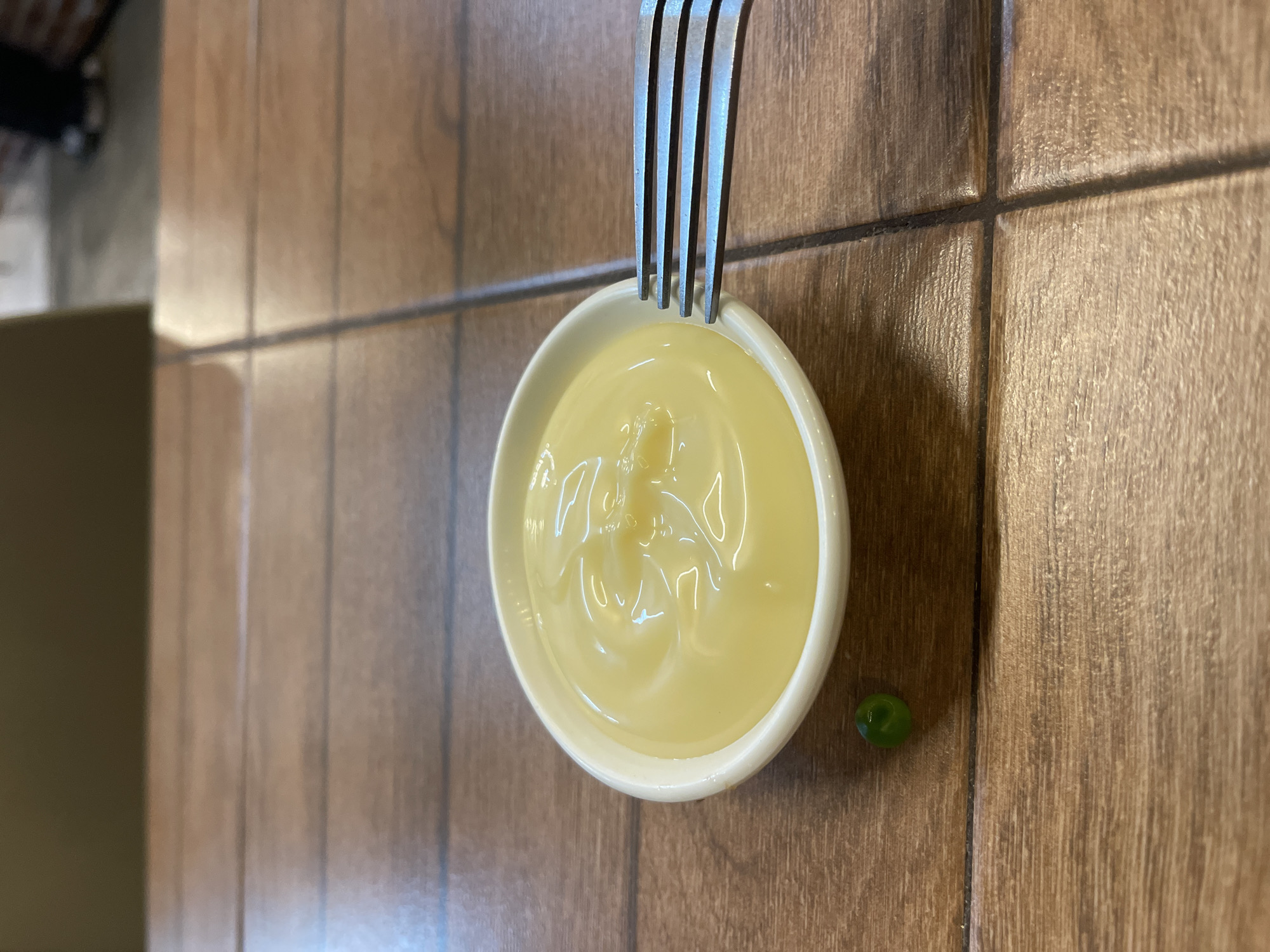A close-up, eye-level shot shows a small white ramekin filled with creamy yellow mayonnaise, positioned on a wooden table. A single green pea rests to the left of the ramekin. A metal fork, with its tines partially submerged in the mayonnaise, is visible to the right of the ramekin. The table surface is composed of wooden planks, with visible grout lines between them. The background is blurred, suggesting an indoor environment, possibly a restaurant or home.