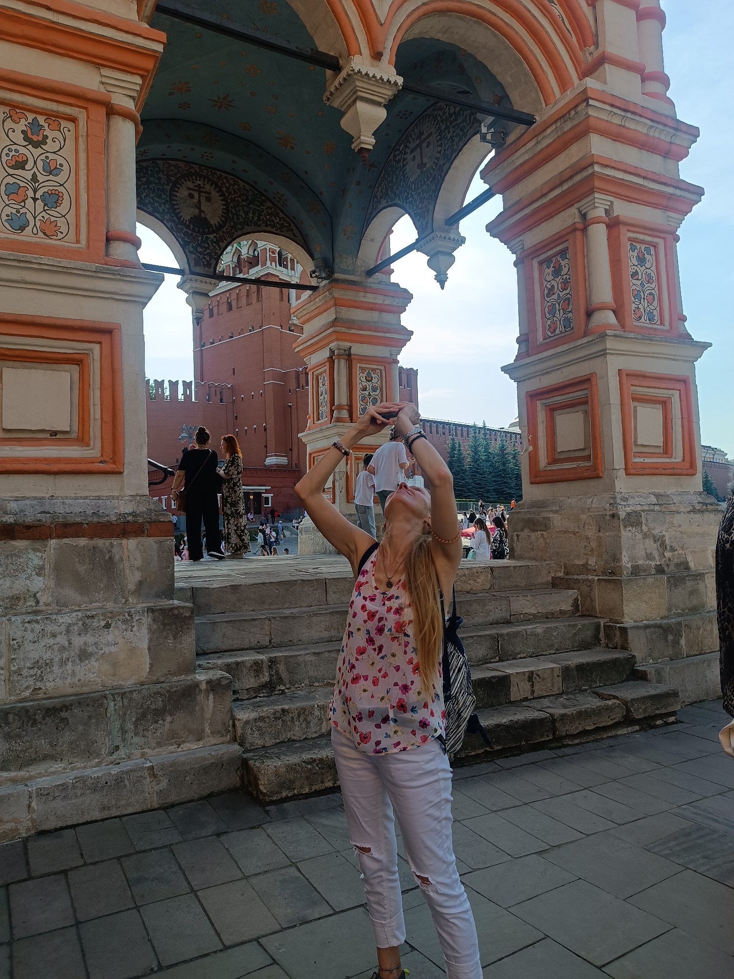 This detailed image captures an outdoor scene in Elektrougli, Russia, featuring a fair-skinned woman in the foreground engaged in photography. She has long blonde hair, styled back, and is wearing a white sleeveless top with a colorful floral pattern of red, pink, blue, and green flowers, paired with white ripped jeans. Bracelets are visible on her left wrist, and a necklace with a small pendant adorns her neck. She holds a dark-colored smartphone with both hands, tilting her head sharply upwards and backward, aiming the device towards the ornate archway directly above her. A dark-colored backpack with a striped pattern is worn on her back. She stands on a paved surface of dark grey square tiles, at the base of worn, light grey stone steps.
The woman is positioned within a large, decorative architectural structure composed of robust, light-colored stone pillars and multiple archways, accented with prominent orange-red borders and details. The inner ceilings of the larger archways are painted light blue with scattered gold and blue floral or star-like patterns. Underneath a smaller arch, a darker, intricate pattern with a central circular motif containing a cross-like symbol is visible. Vertical panels on the pillars display vibrant floral designs in red, blue, and green within swirling vine motifs, enclosed in square frames. Other panels feature plain orange-bordered squares. Dark, horizontal metal support bars extend between the pillars.
In the mid-ground and background, visible through the archways, are significant red brick walls of a fortress-like structure, featuring battlements along their top edge. A distinct red brick tower with a green-domed roof is partially visible through one archway. Several dark green coniferous trees stand behind the red walls. Numerous other people are scattered throughout the background, walking and standing, indicating a public and active environment. The sky above is clear and light blue, suggesting bright, fair weather conditions.