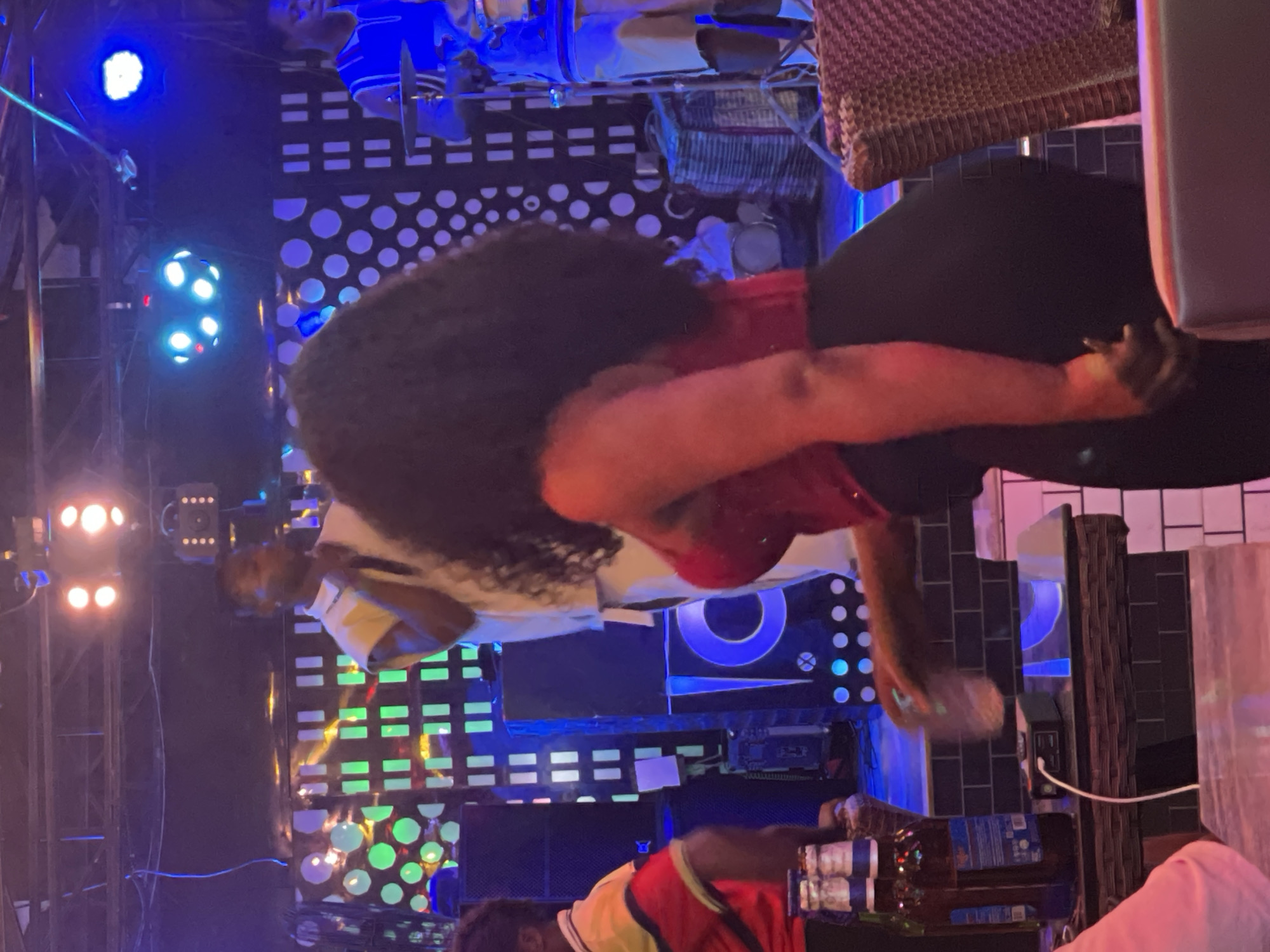 The image depicts an indoor live music event at an entertainment venue in Obosi, Nigeria. The scene is characterized by dim ambient lighting accentuated by numerous colorful stage and decorative lights, casting hues of blue, green, red, and white.
On a raised stage in the mid-ground, a male vocalist wearing a light-colored, sleeveless top is performing, holding a microphone. To his left, another individual in a red, yellow, and blue collared shirt is partially visible, facing towards the stage's center. To the vocalist's right, a male drummer in a blue sleeveless top is positioned behind a drum set. Large black speaker cabinets, some with blue illuminated circular and horizontal accents, are present on the stage. The background wall behind the stage features illuminated geometric patterns composed of squares and circles.
In the foreground, a woman with long, dark, curly hair, dressed in a red top and black bottoms, stands partially facing away from the camera, holding a clear glass in her right hand. On a woven-patterned table to her left, two glass bottles with blue labels and a small black electronic device connected by a white cable are visible. A portion of a woven chair back is present in the far right foreground. The environment indicates an active, social gathering focused on the live performance.