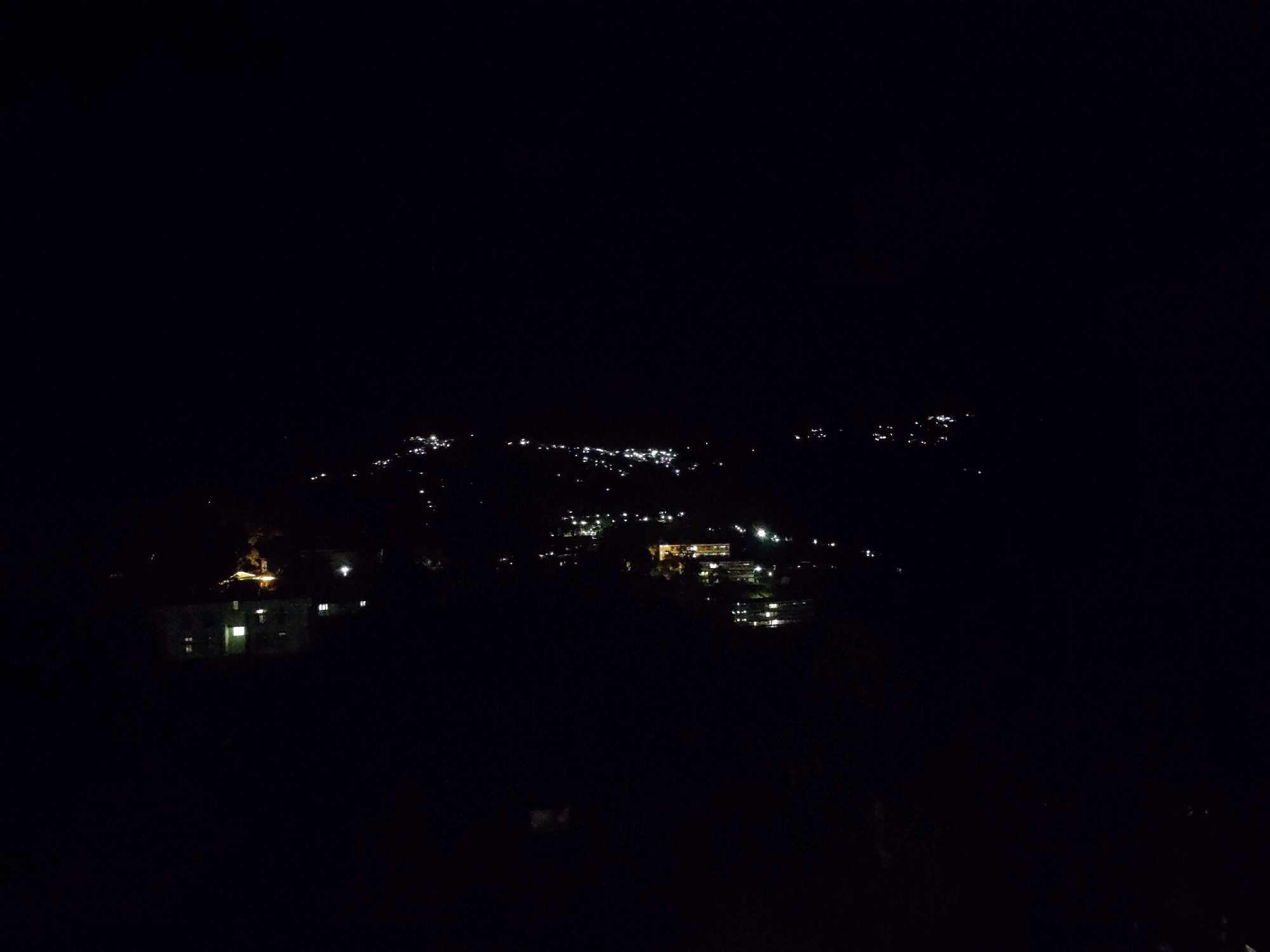An elevated, wide shot captures a dark night sky over a dimly lit settlement in Mokokchung, India. The landscape is dominated by the blackness of night, punctuated by clusters of artificial lights emanating from buildings and streetlights scattered across the terrain. In the foreground, a dark, silhouetted structure, possibly a residential building, is visible on the left, with a few illuminated windows offering a faint glow. Further into the distance, more lights form a hazy constellation, suggesting a spread of habitation. The terrain appears uneven, with the lights following the contours of hills or valleys. No people are visible in the frame. The overall impression is one of a quiet, remote settlement at night.