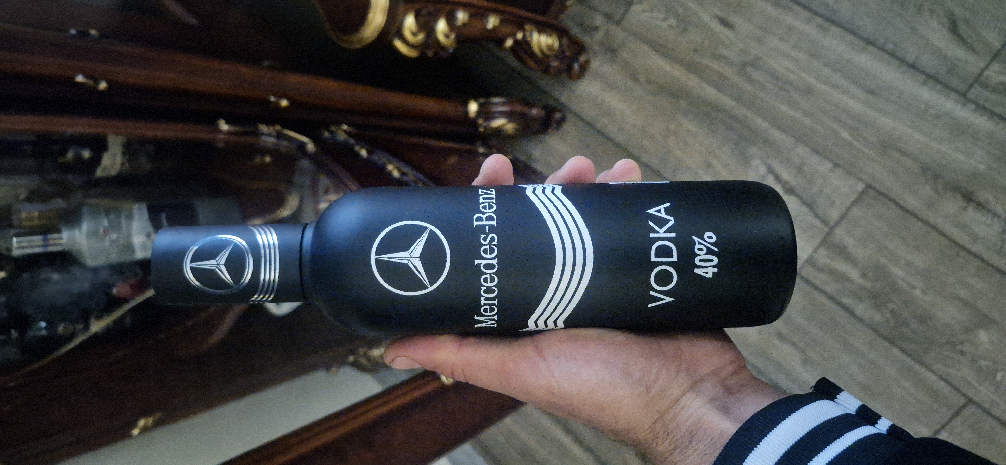A hand holds a bottle of Mercedes-Benz vodka, labeled "40%," against the backdrop of a wooden table with ornate carvings and a section of gray flooring. The location is Yerevan, Armenia. The bottle is black with white lettering and Mercedes-Benz branding. The hand holding the bottle has short fingernails and some visible veins. A striped sleeve is visible on the arm of the person holding the bottle. Other bottles are visible in the background on the table.
